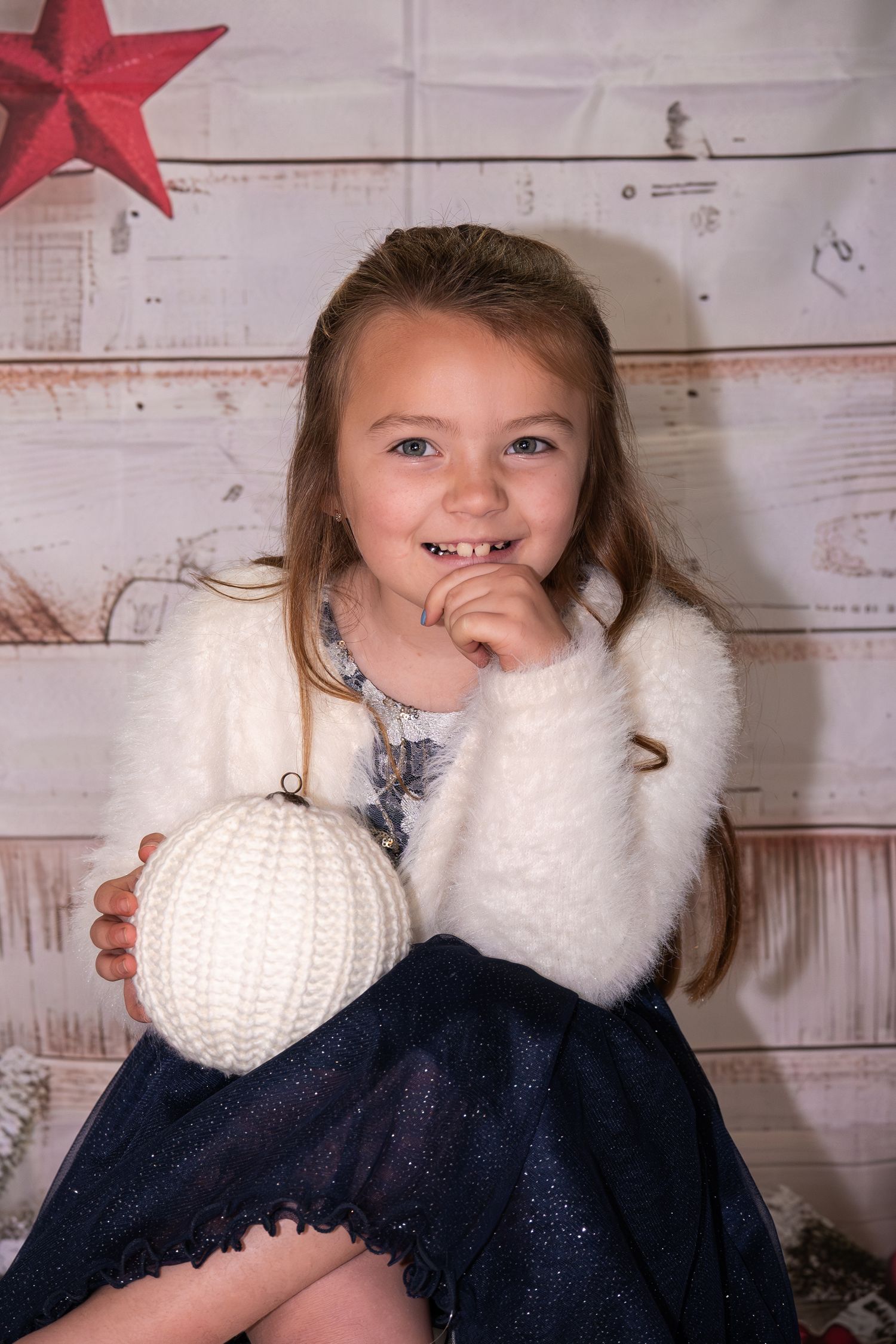 Child in navy sweater with white fur collar sits by red star decoration against rustic white wooden wall.