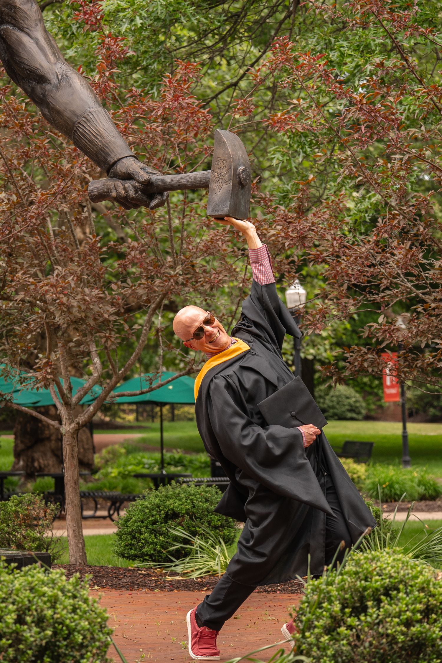 Graduate in black robe and red shoes poses playfully near flowering trees.
