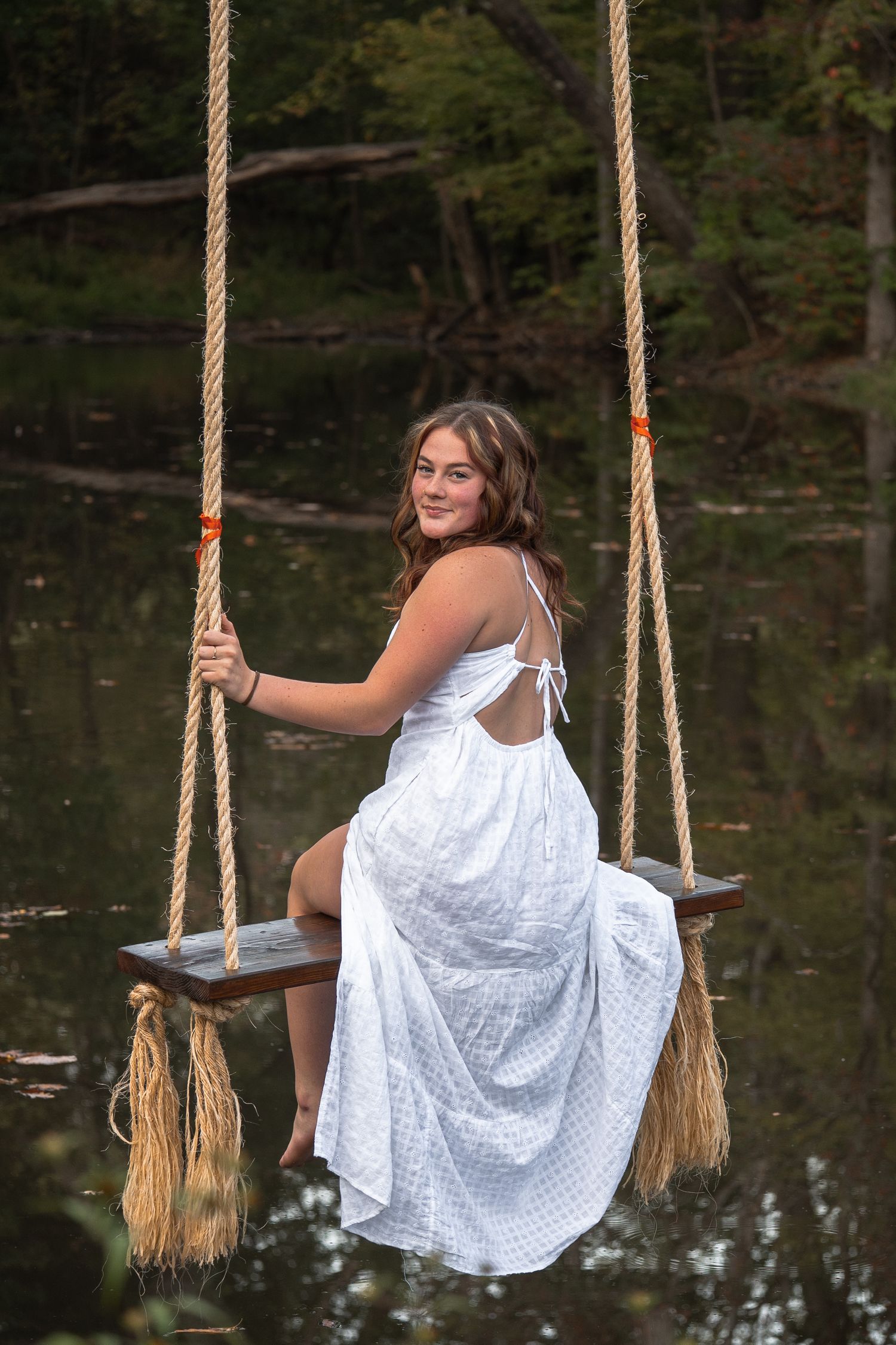 A person in a flowing white dress swings gracefully on a rope over a reflective pond in a wooded setting.
