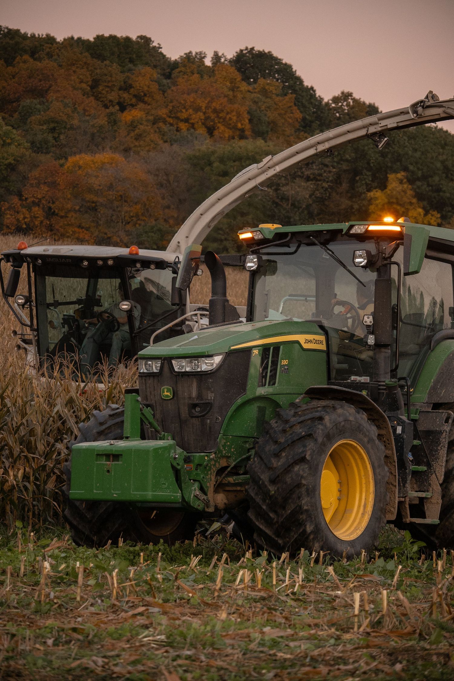 Line of John Deere tractors harvesting in a cornfield at sunset.