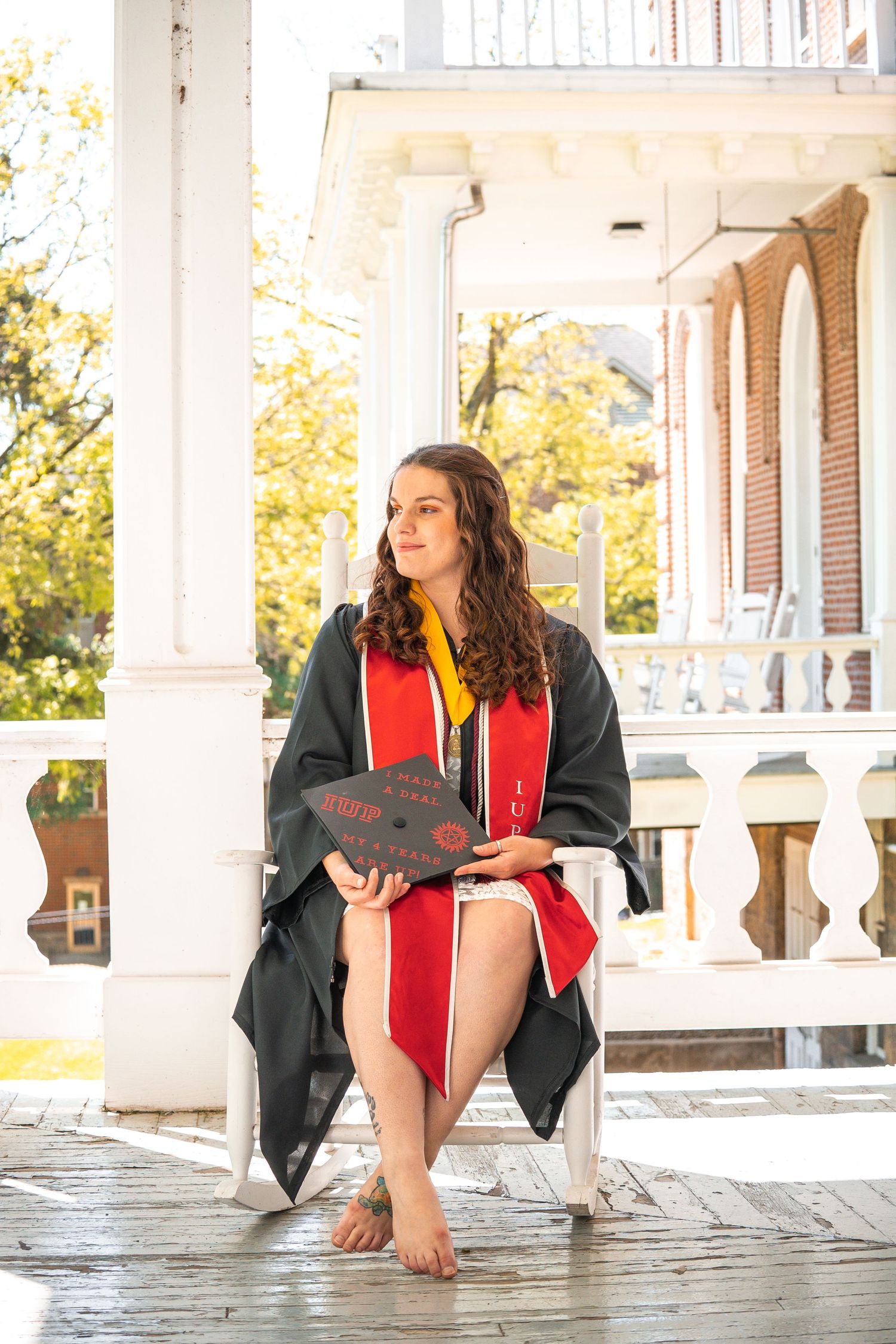 A graduate in cap and gown sits on a white porch railing during a photo session.