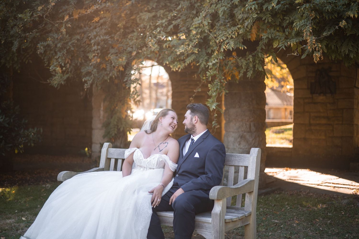 A romantic wedding couple shares intimate moments on a bench in a garden setting at sunset.