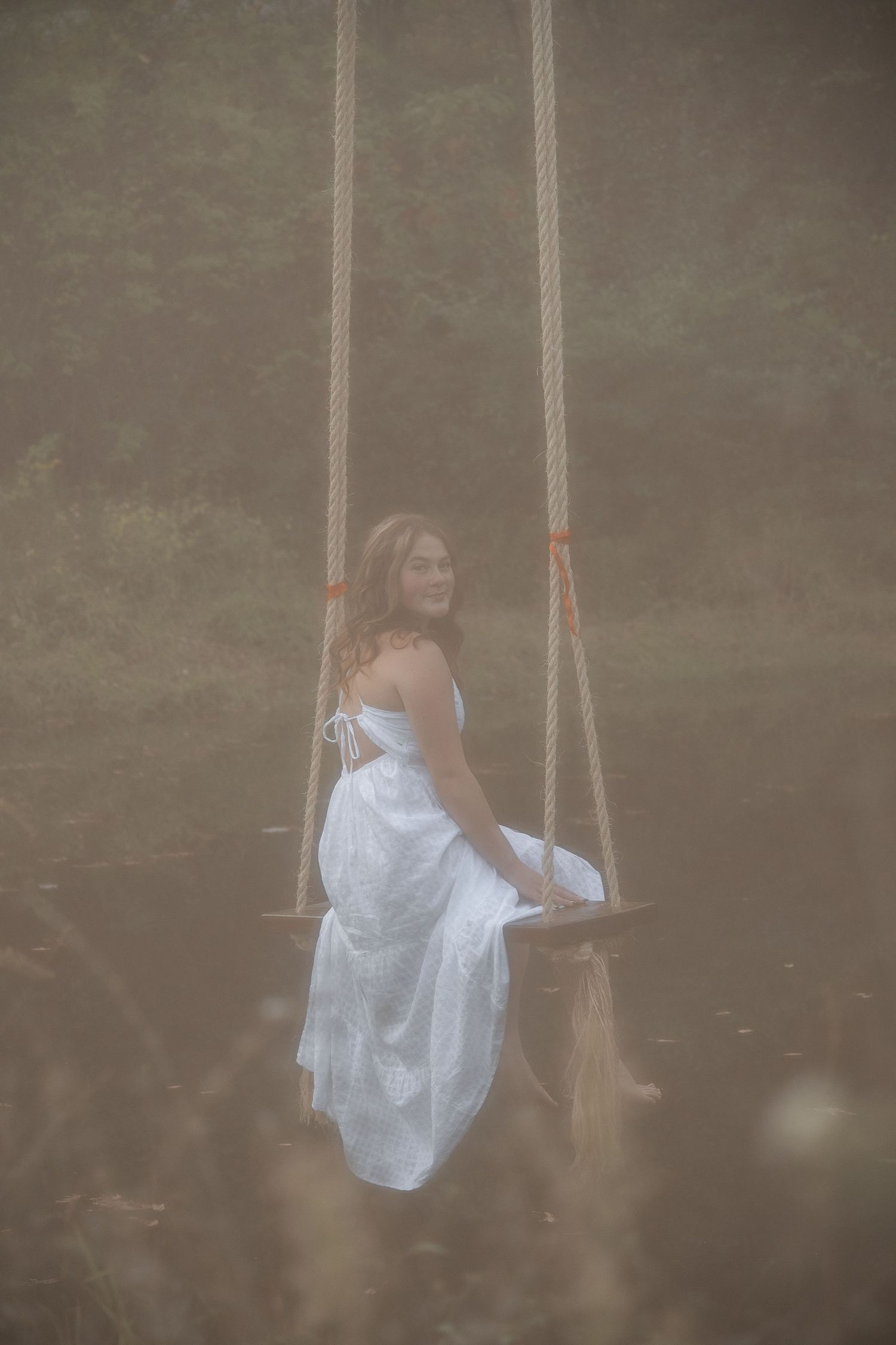 A dreamy ethereal scene shows someone in a flowing white dress sitting on a rope swing in a misty outdoor setting.