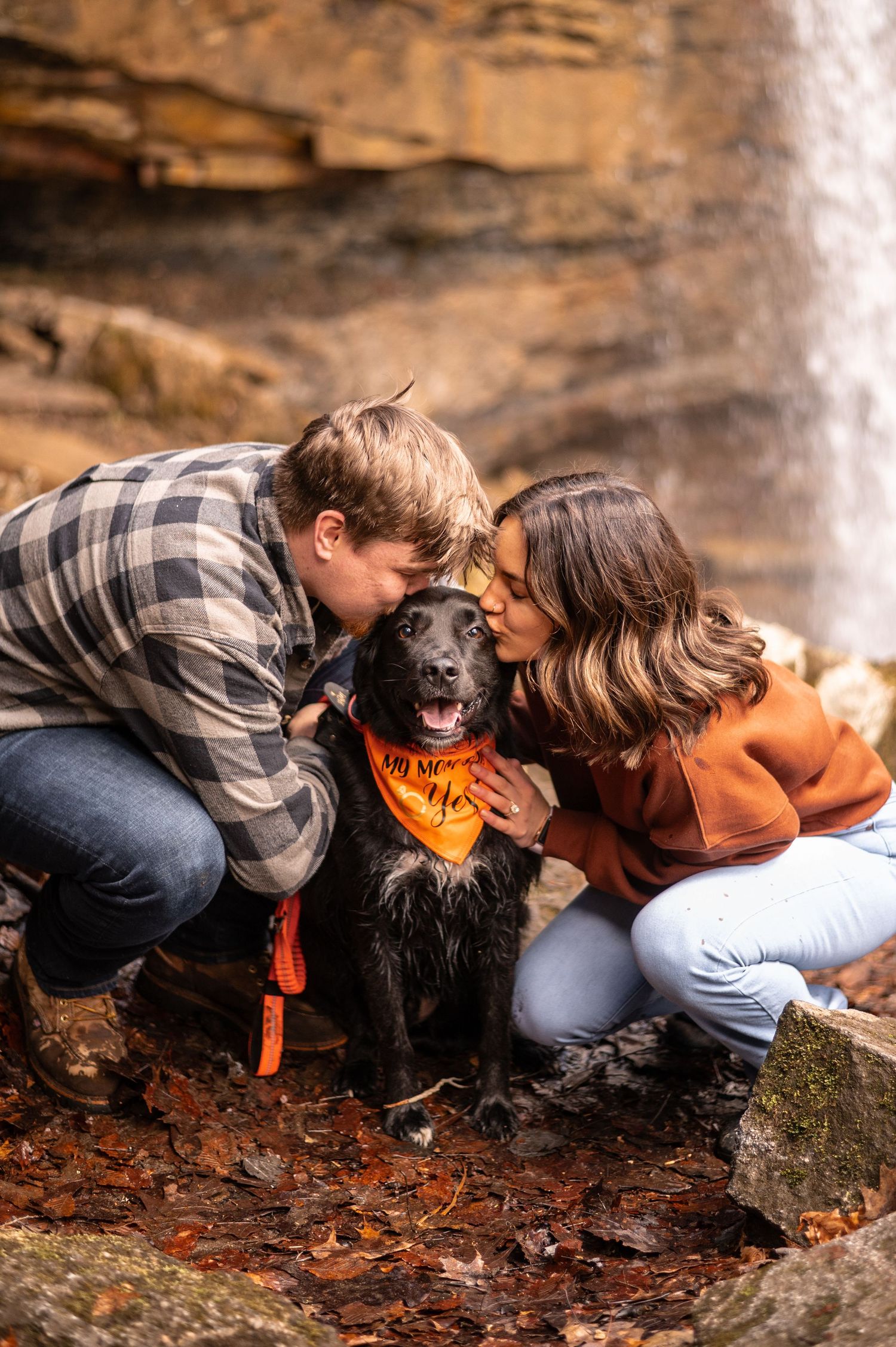 A couple and their black dog share a tender moment among fallen autumn leaves near a waterfall.