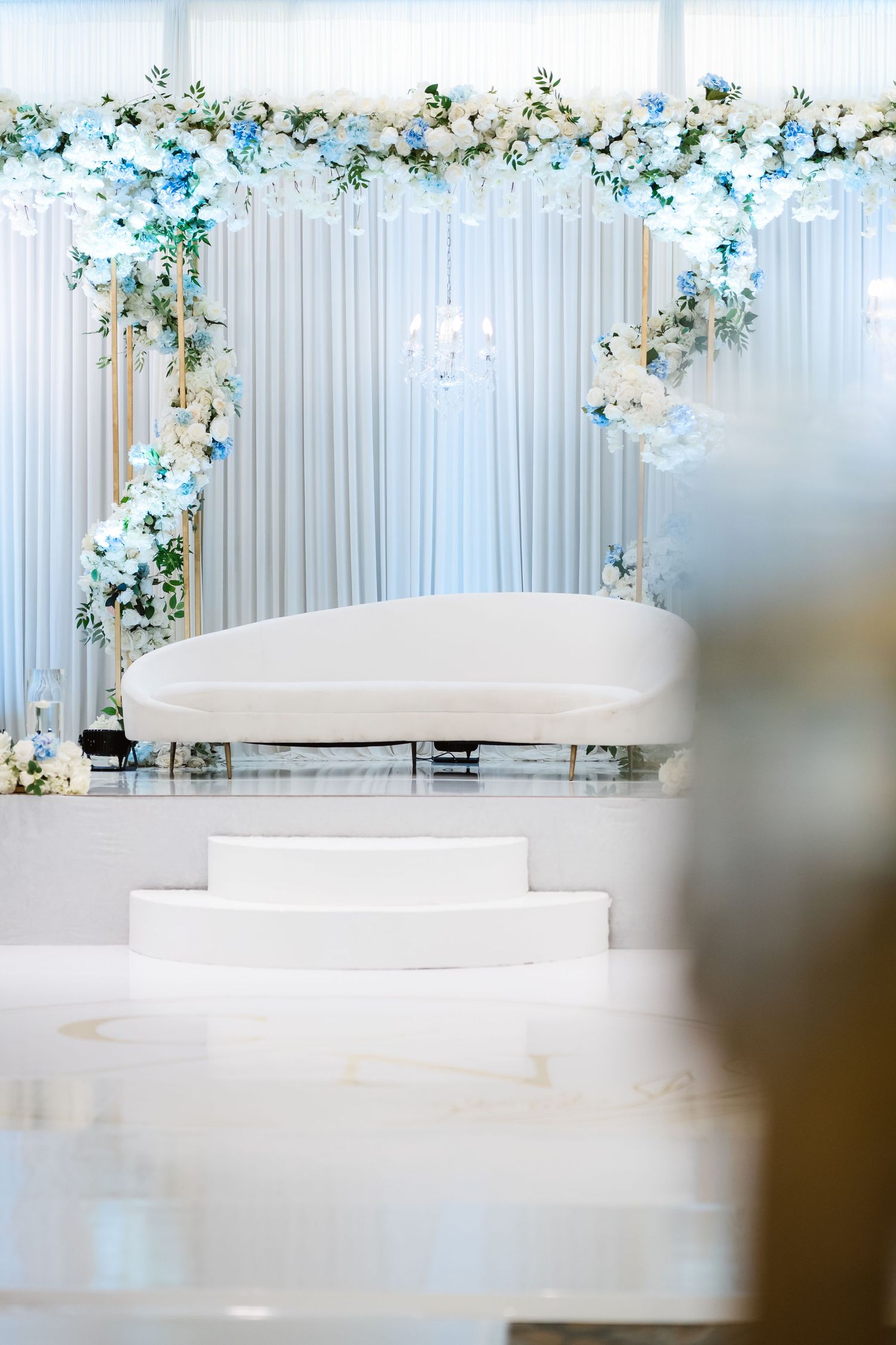 Elegant white wedding backdrop with blue and white floral arrangements and draped fabric.