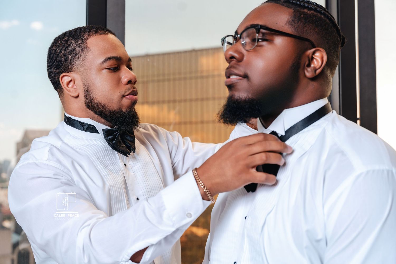 Two men in white dress shirts help each other adjust their bow ties.
