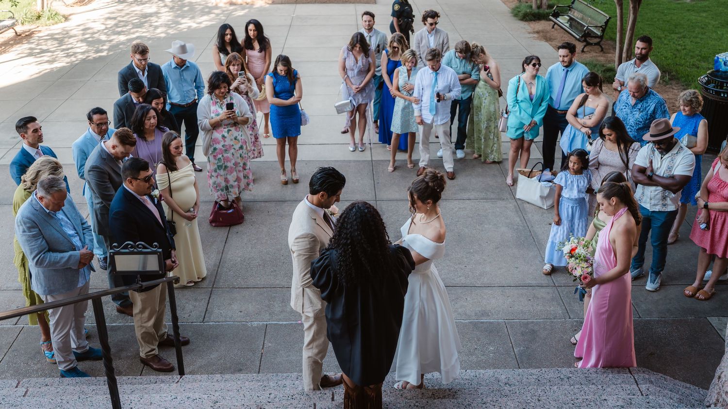 A large wedding party gathers for a panoramic group photo on a sunny day outdoors.