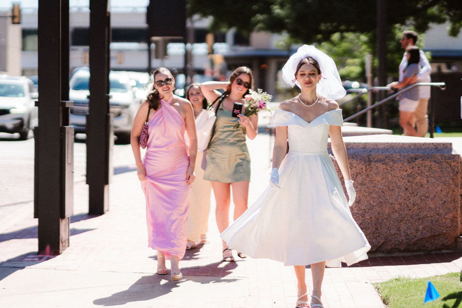 A group walks down a sunny outdoor path at a wedding celebration with pink and white dresses.