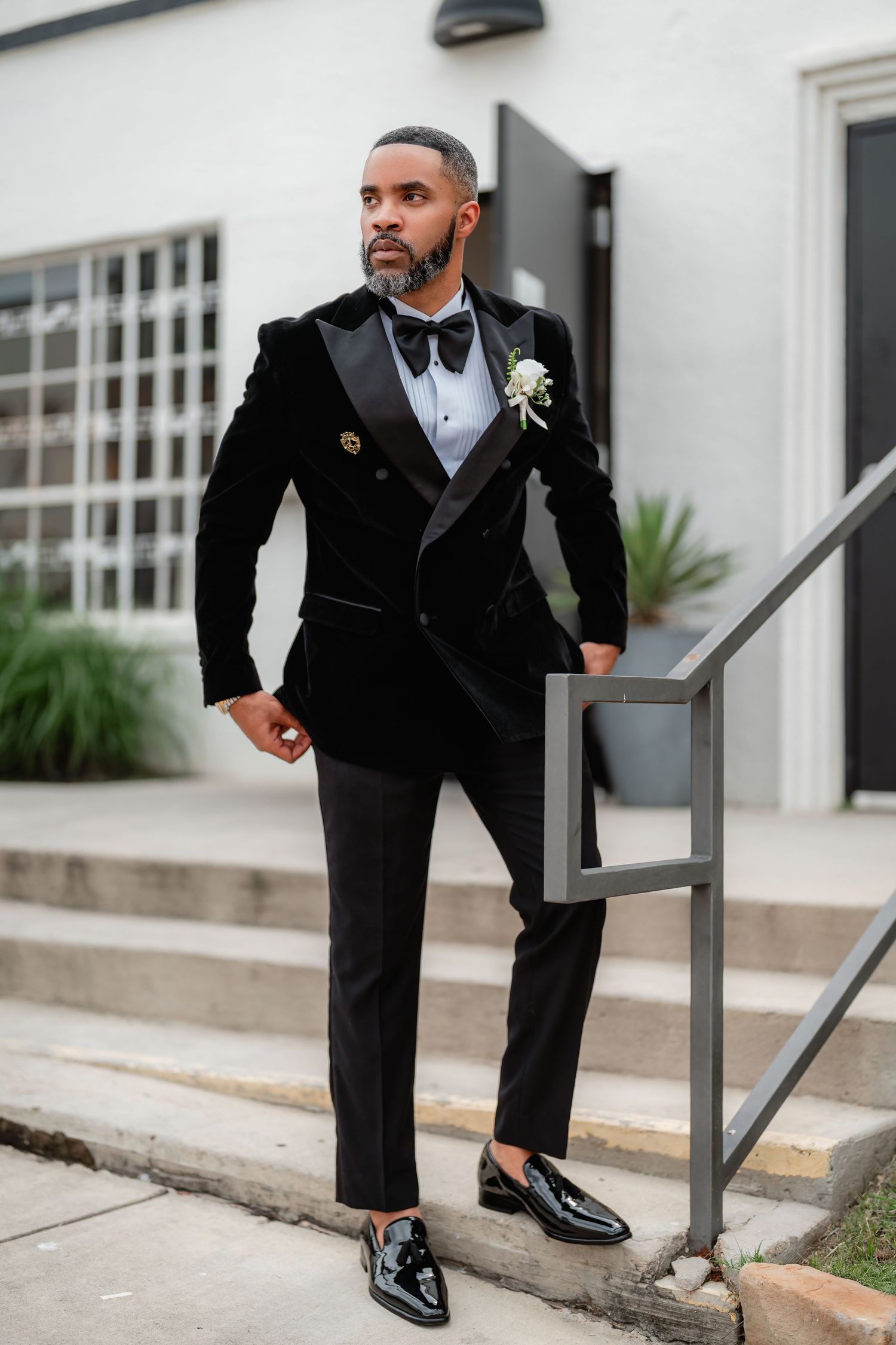 Groom in black tuxedo and bow tie poses confidently on modern steps with shiny dress shoes.