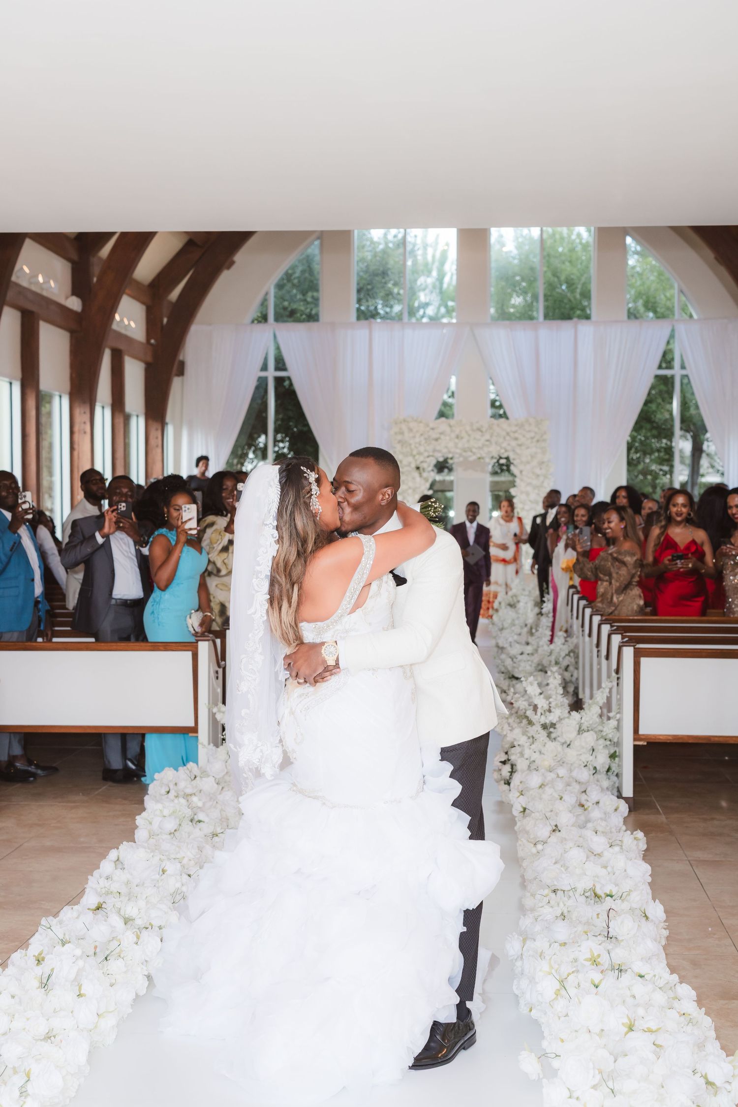 A couple shares their first kiss as newlyweds in a chapel decorated with white floral arrangements along the aisle.