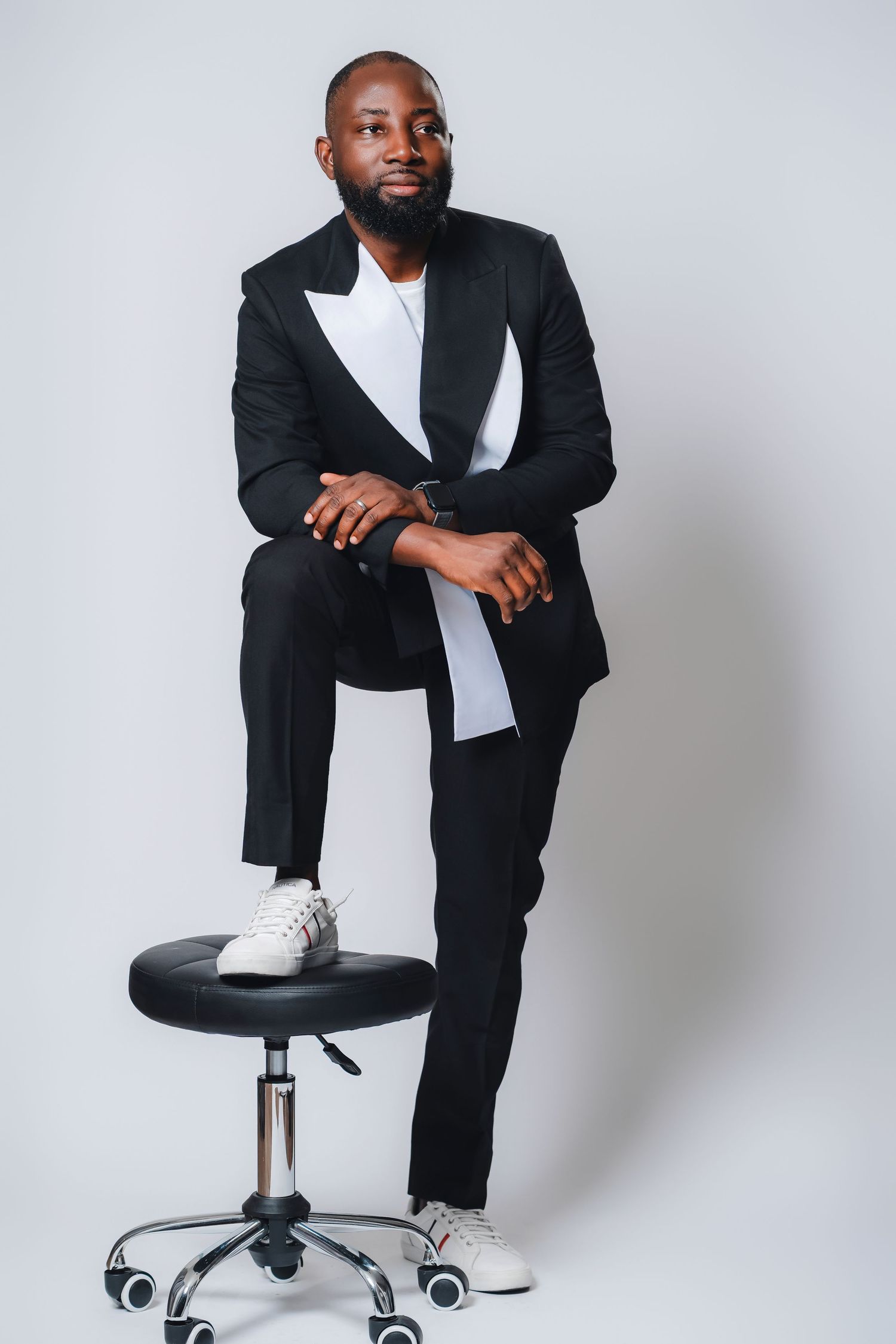 Stylish man in black suit and white sneakers poses confidently on an office chair against light background.