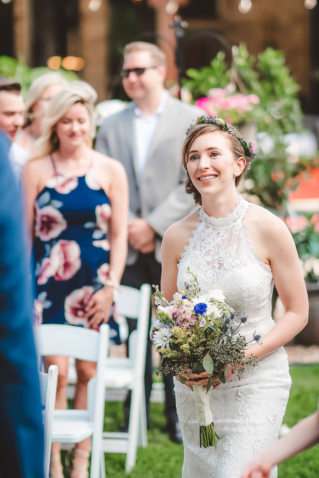 A bride in a lace halter dress walks down the aisle holding a wildflower bouquet.