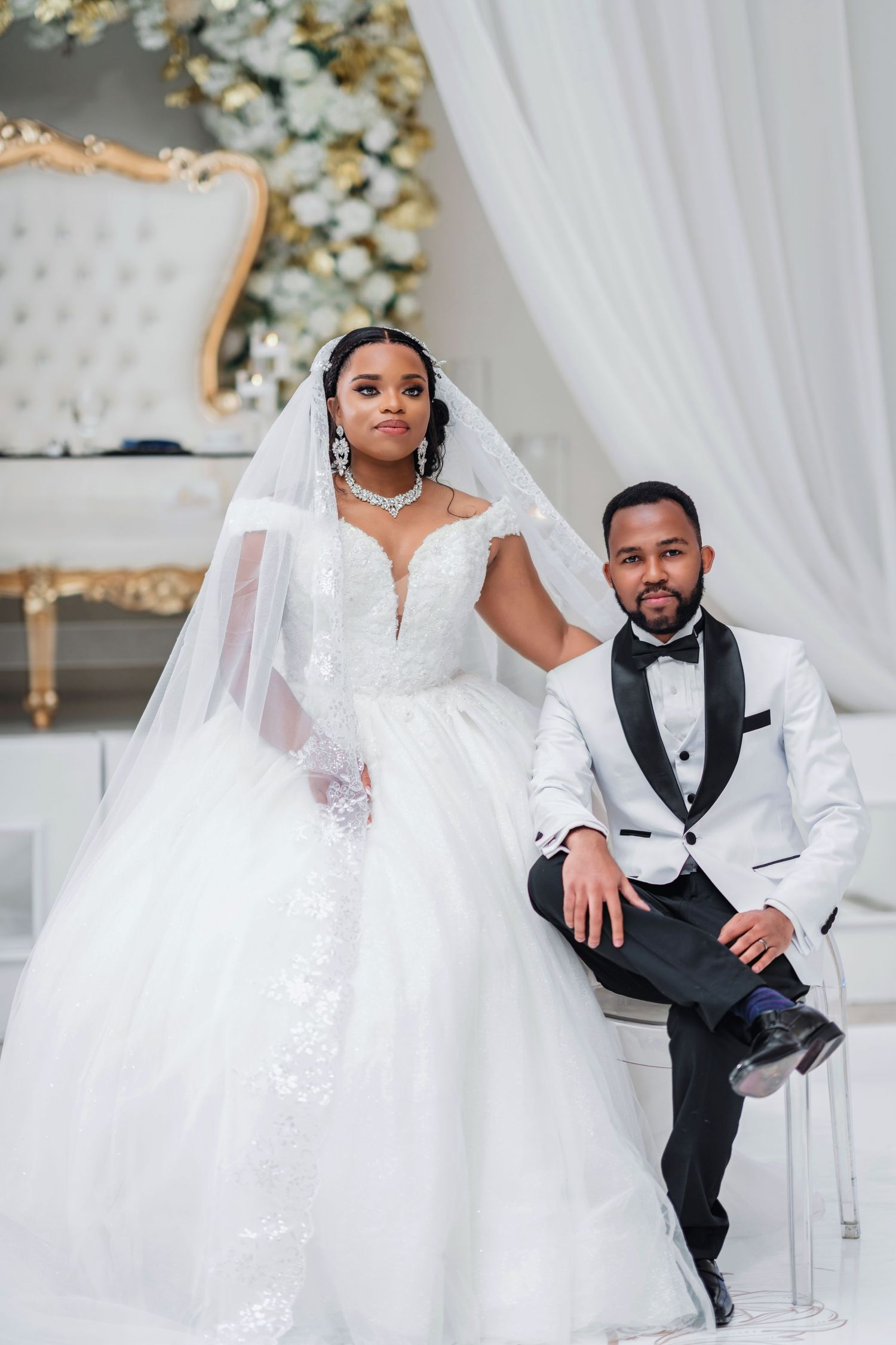 Wedding couple in white and black formal attire seated in elegant chairs.
