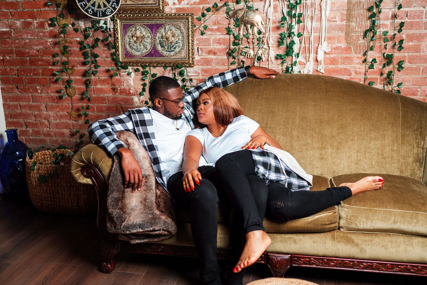 A couple relaxes together on a vintage couch in a cozy brick-walled room with hanging plants.