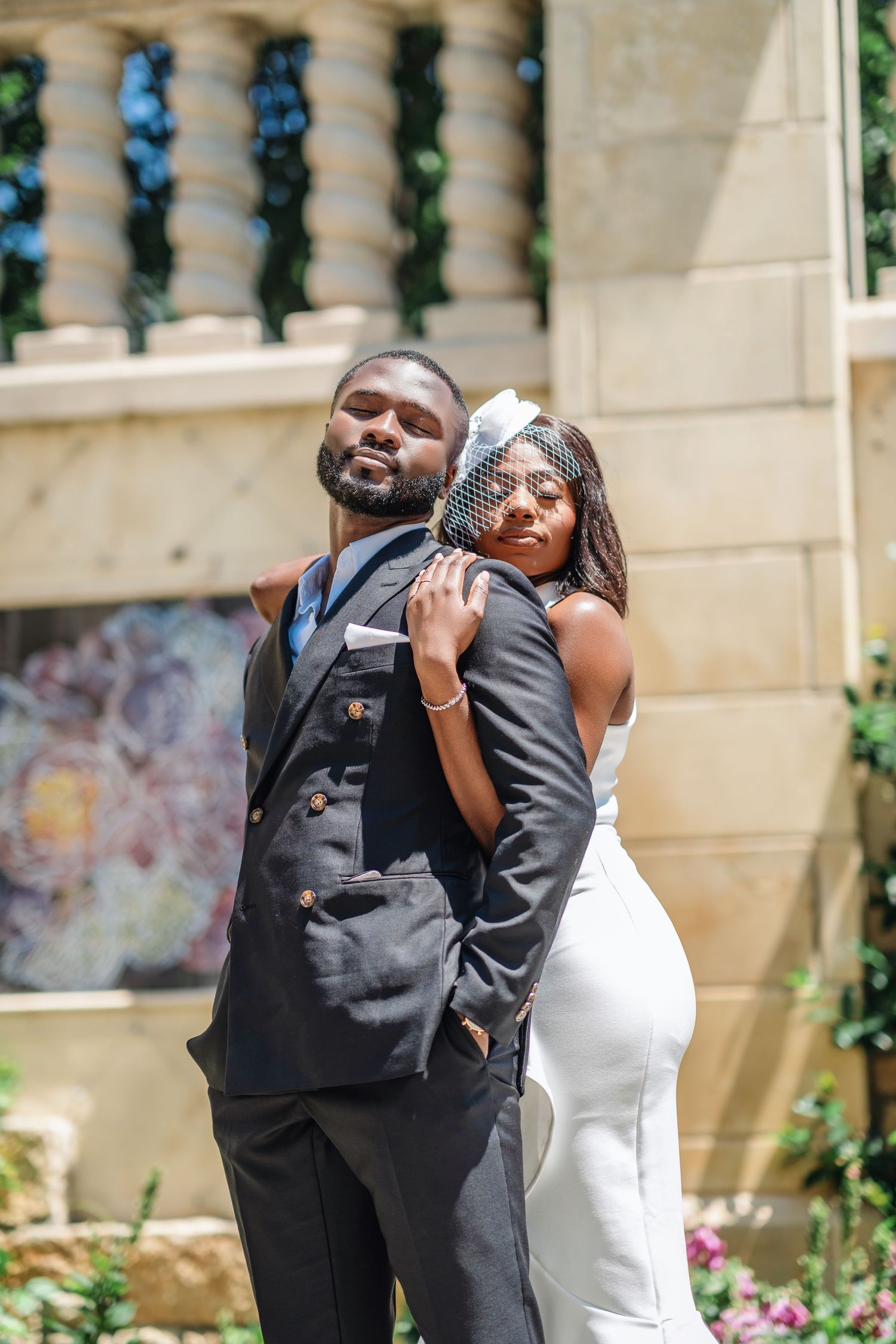 A newlywed couple poses playfully against a stone balustrade in a garden setting.