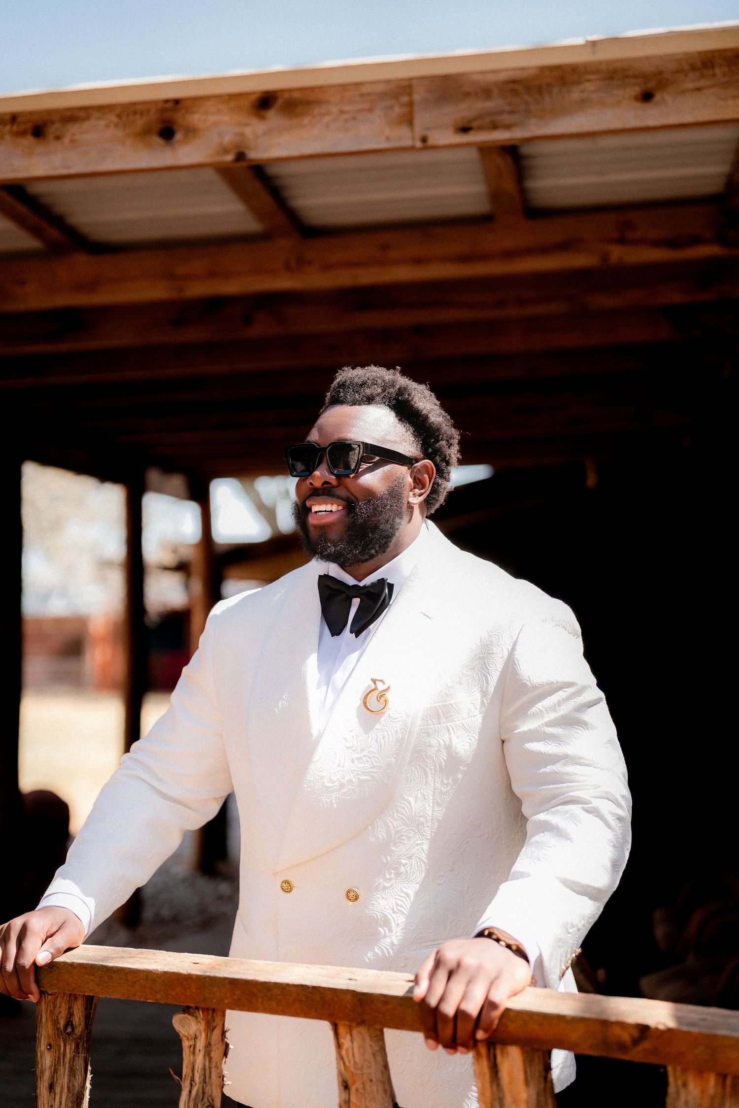 A person in a crisp white tuxedo jacket and bow tie smiles while standing on a rustic wooden porch.