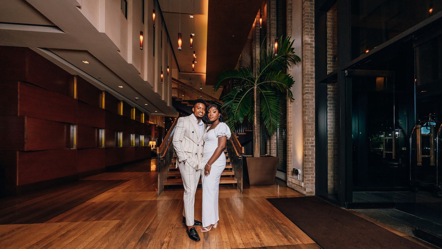 A couple walks hand in hand down an elegantly lit indoor corridor during an evening photoshoot.