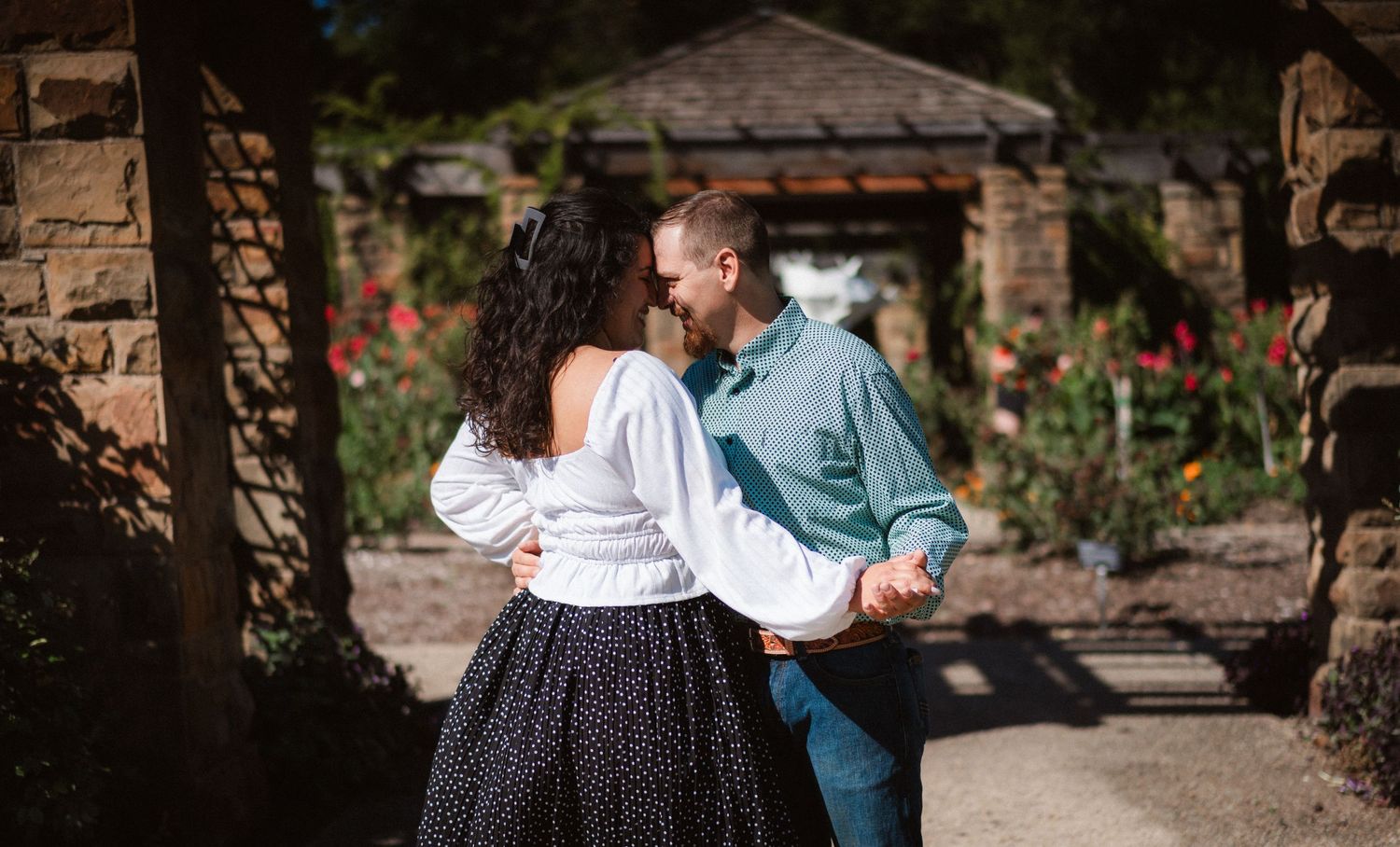 Couple strolls through a scenic garden pathway with stone walls and lush greenery on a sunny day.