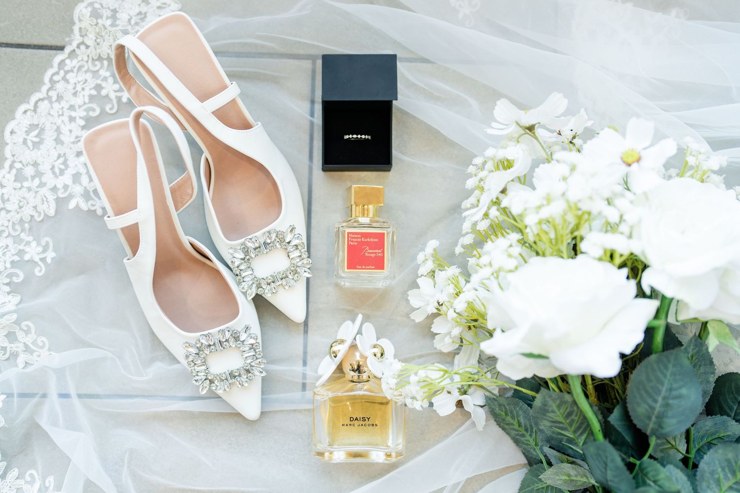 Wedding accessories including white shoes, perfume bottle, and floral decorations arranged on light surface.