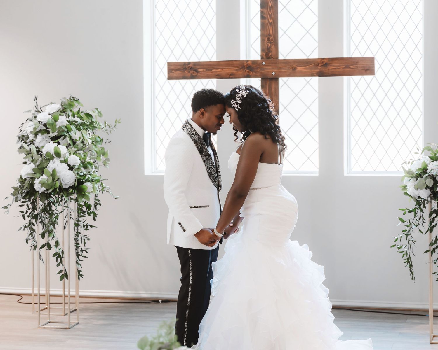 Elegant wedding couple poses in bright white studio space with greenery and cross decor.