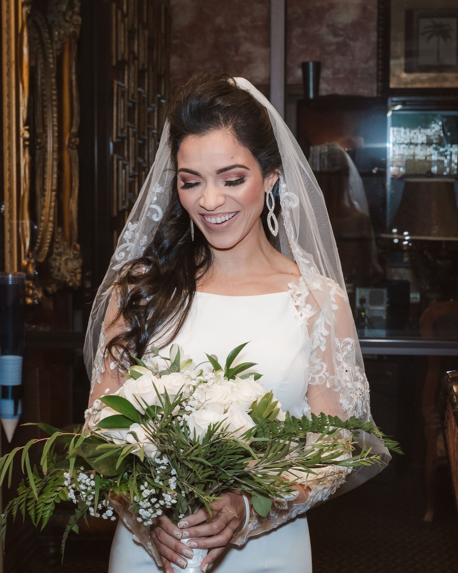A bride in a lace sleeve wedding dress holds a lush greenery bouquet in an elegant indoor setting.