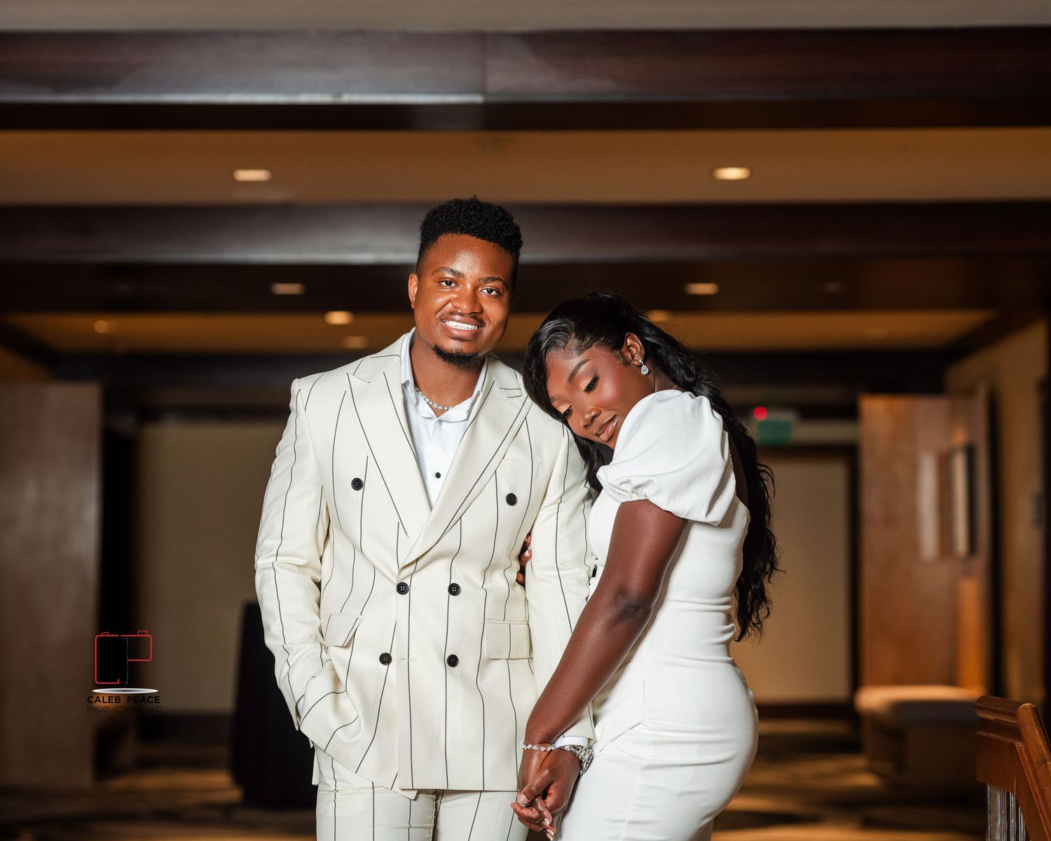 A couple in coordinated white formal attire pose together in a warmly lit indoor hallway.