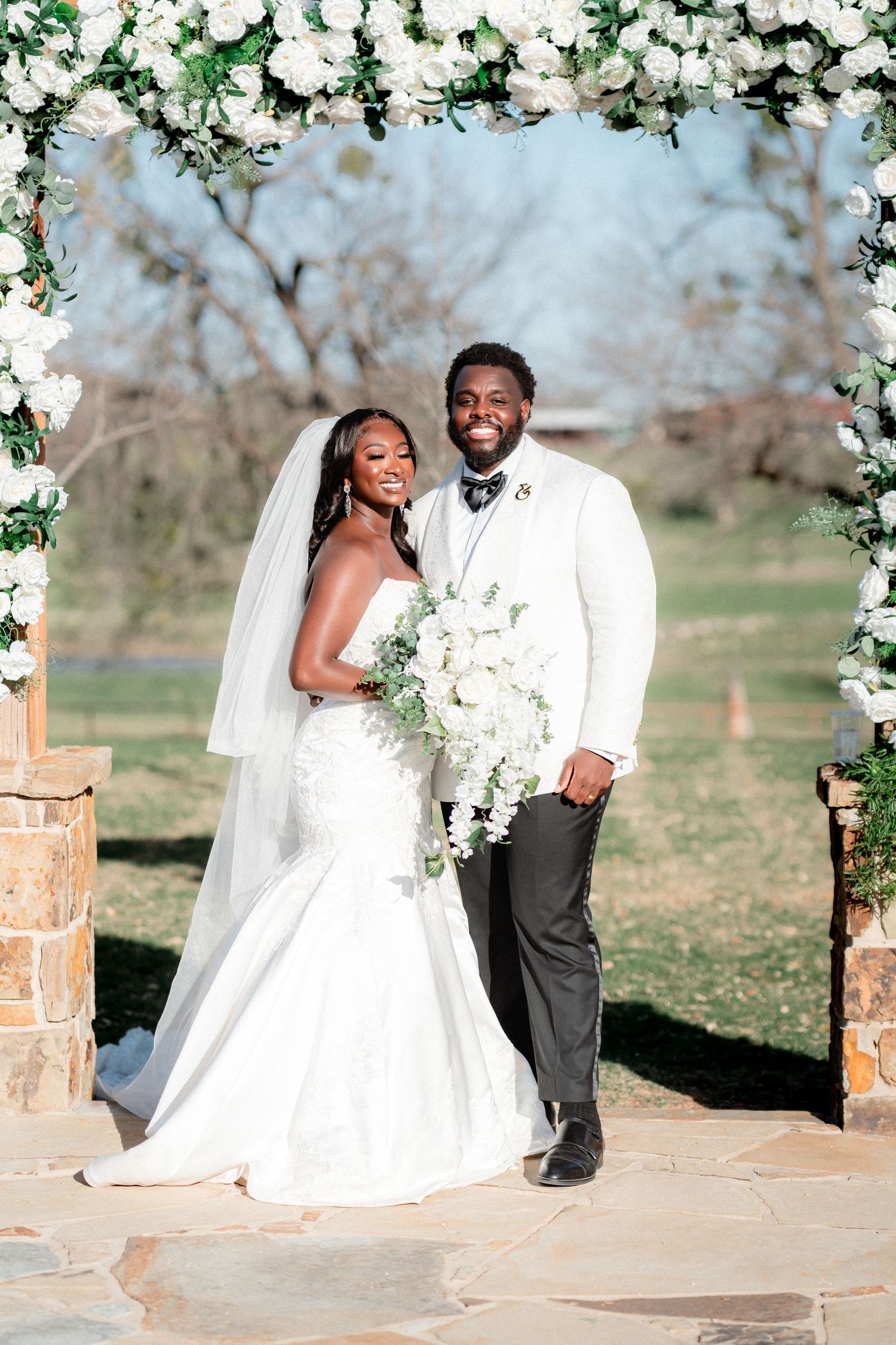 Wedding ceremony setup with white floral arch and happy couple outdoors on a sunny day.