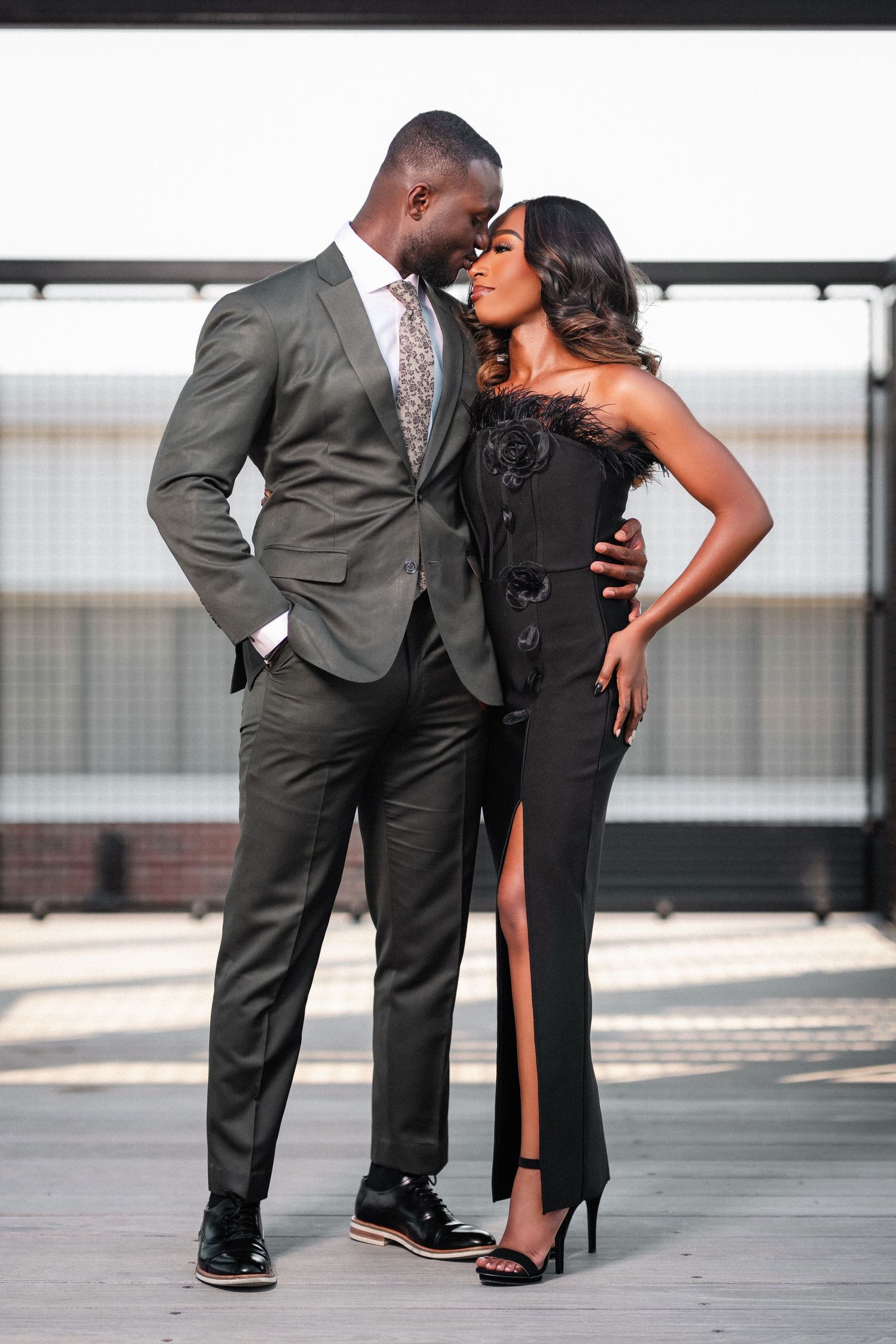 A stylish couple poses together in formal attire with a grey suit and black evening gown against an urban backdrop.