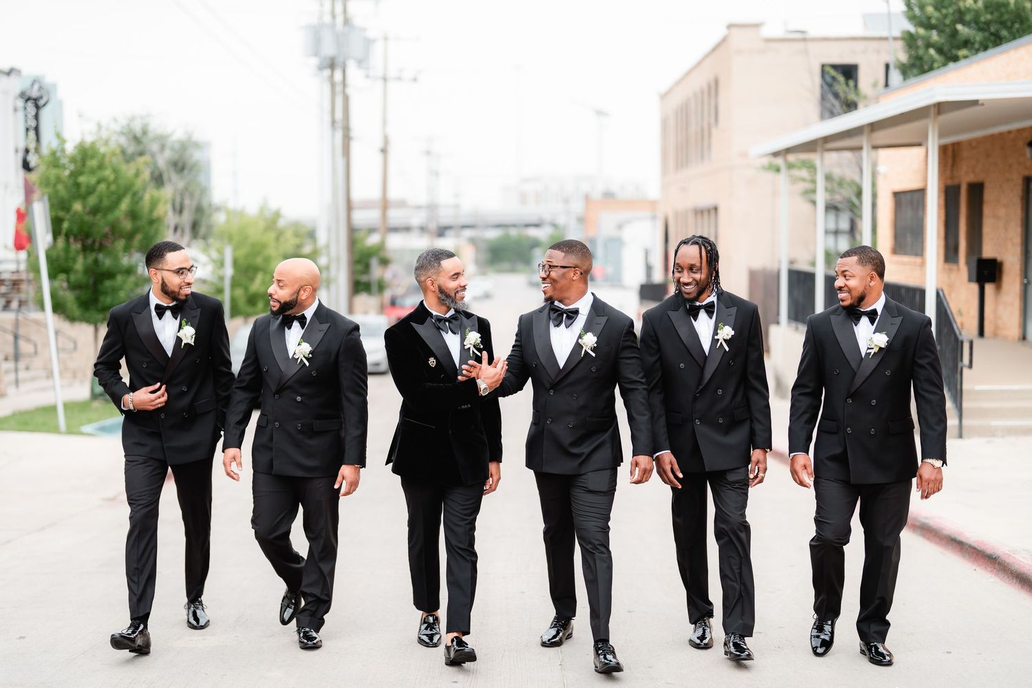 Wedding party in formal black attire walking in line along sidewalk outside brick building.