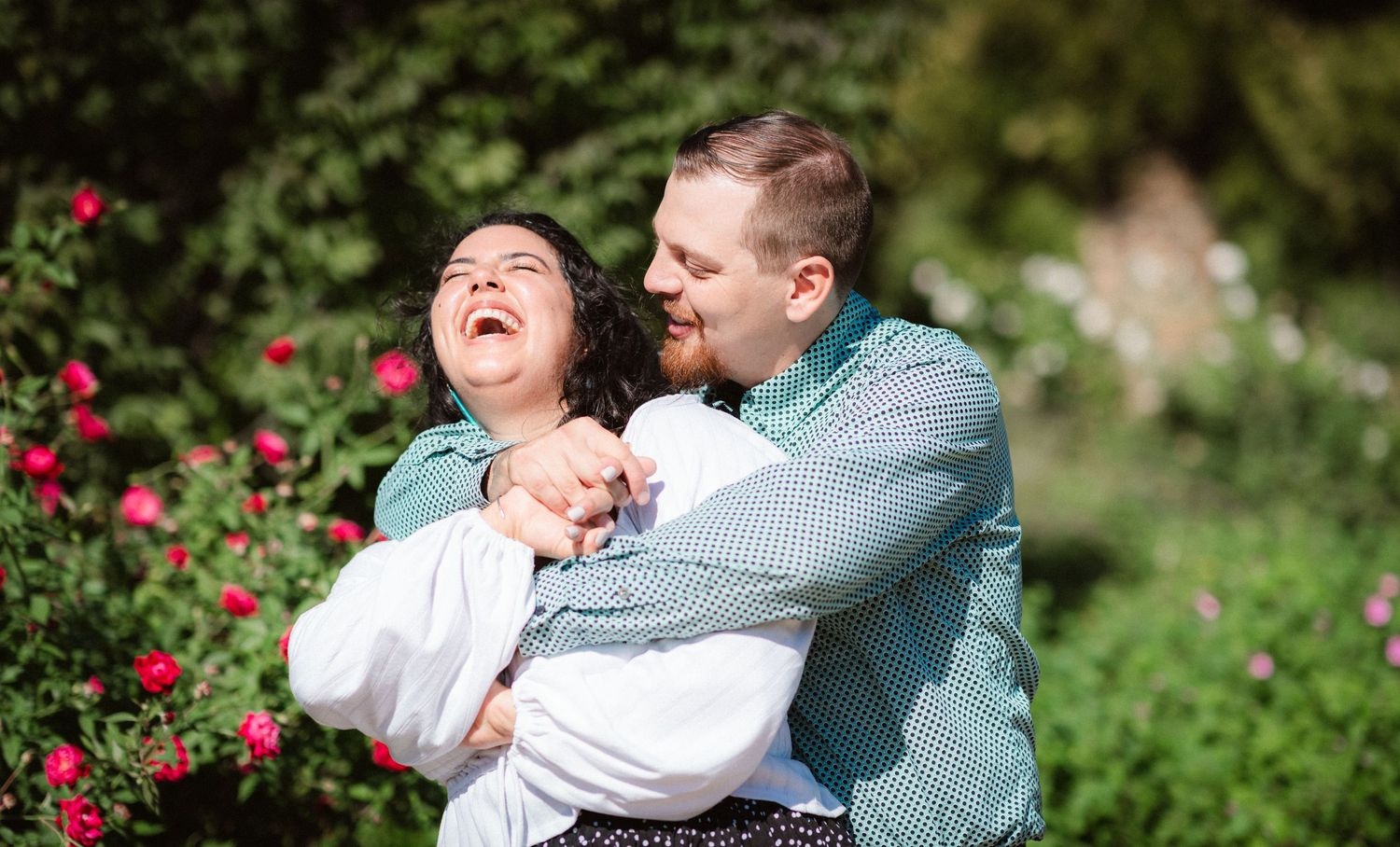 A couple embraces and laughs together in a lush garden setting with red flowers.