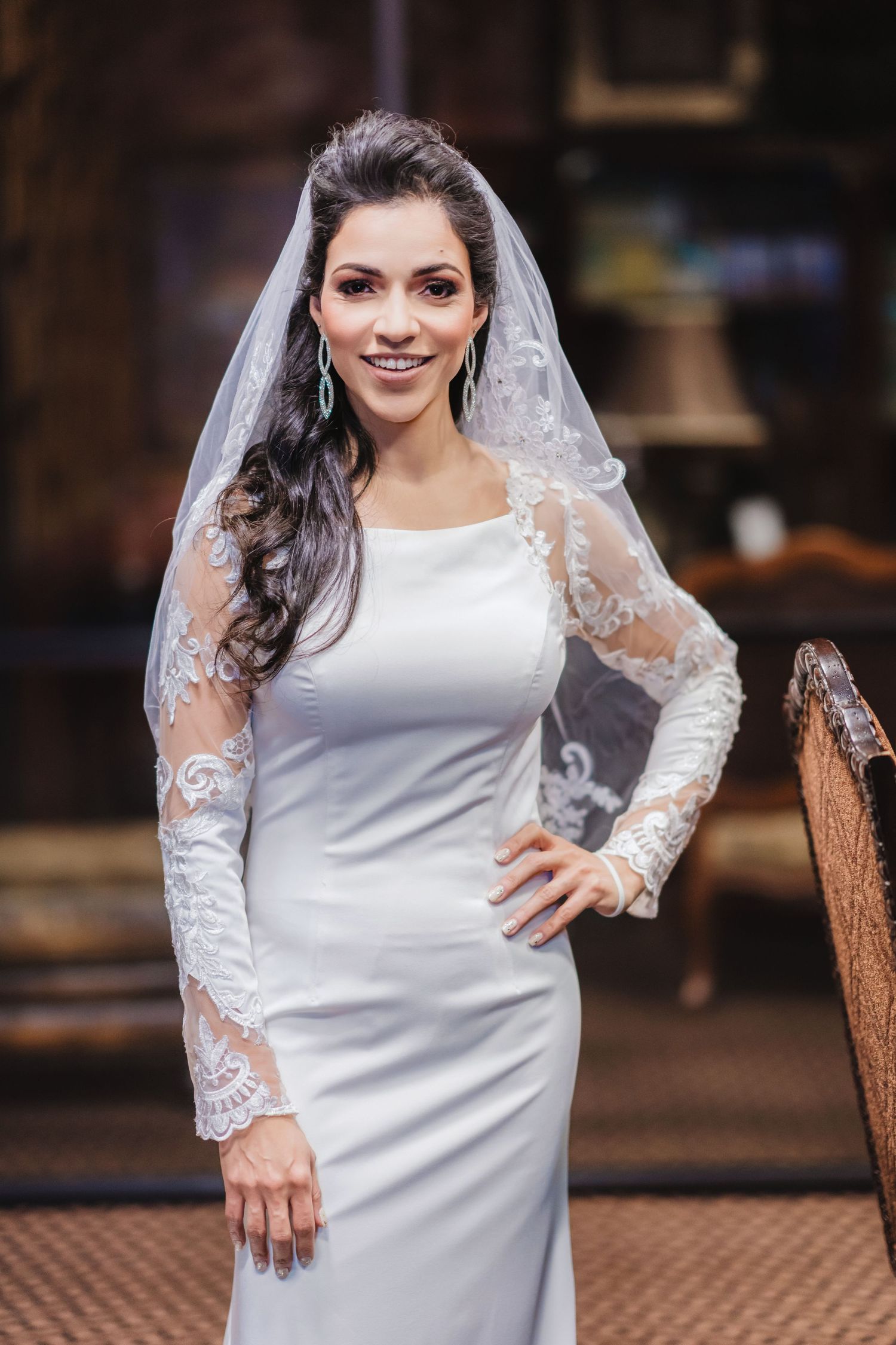 A bride in a white lace-sleeved wedding dress poses elegantly on an ornate chair.
