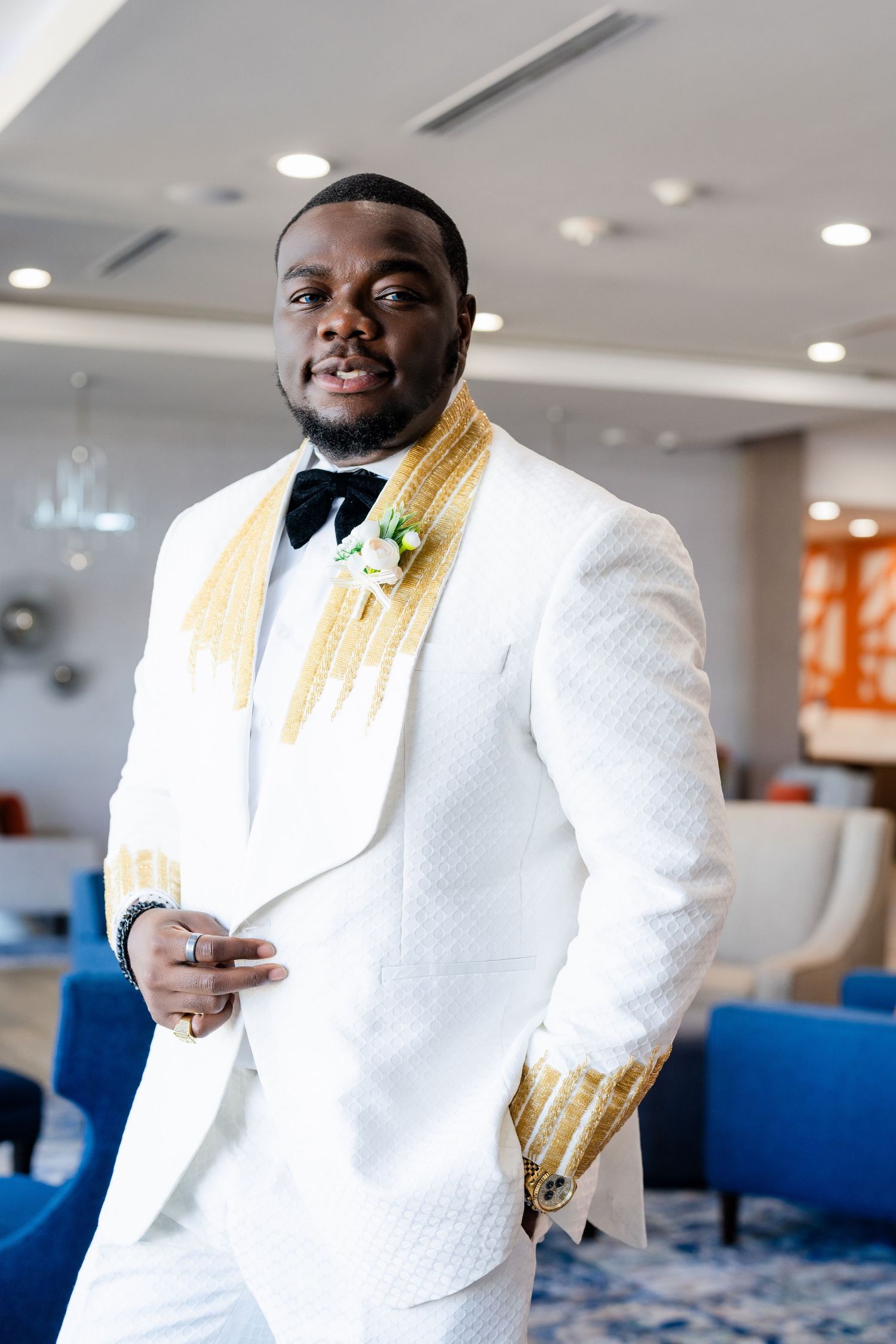 Groom wearing crisp white tuxedo jacket with gold accents and black bow tie poses in elegant indoor setting.