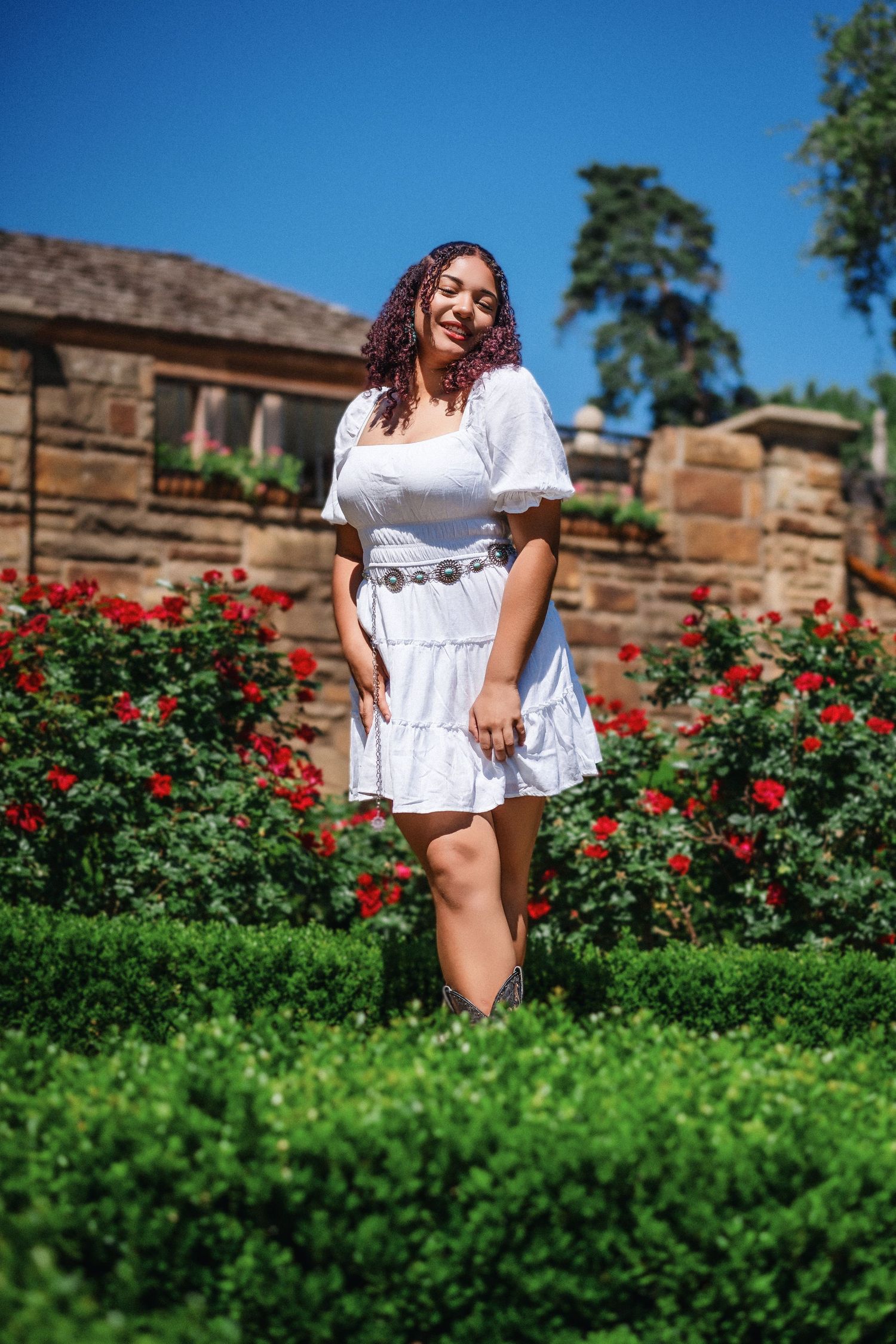 A person in a white summer dress poses in a garden with vibrant red roses and green hedges on a sunny day.