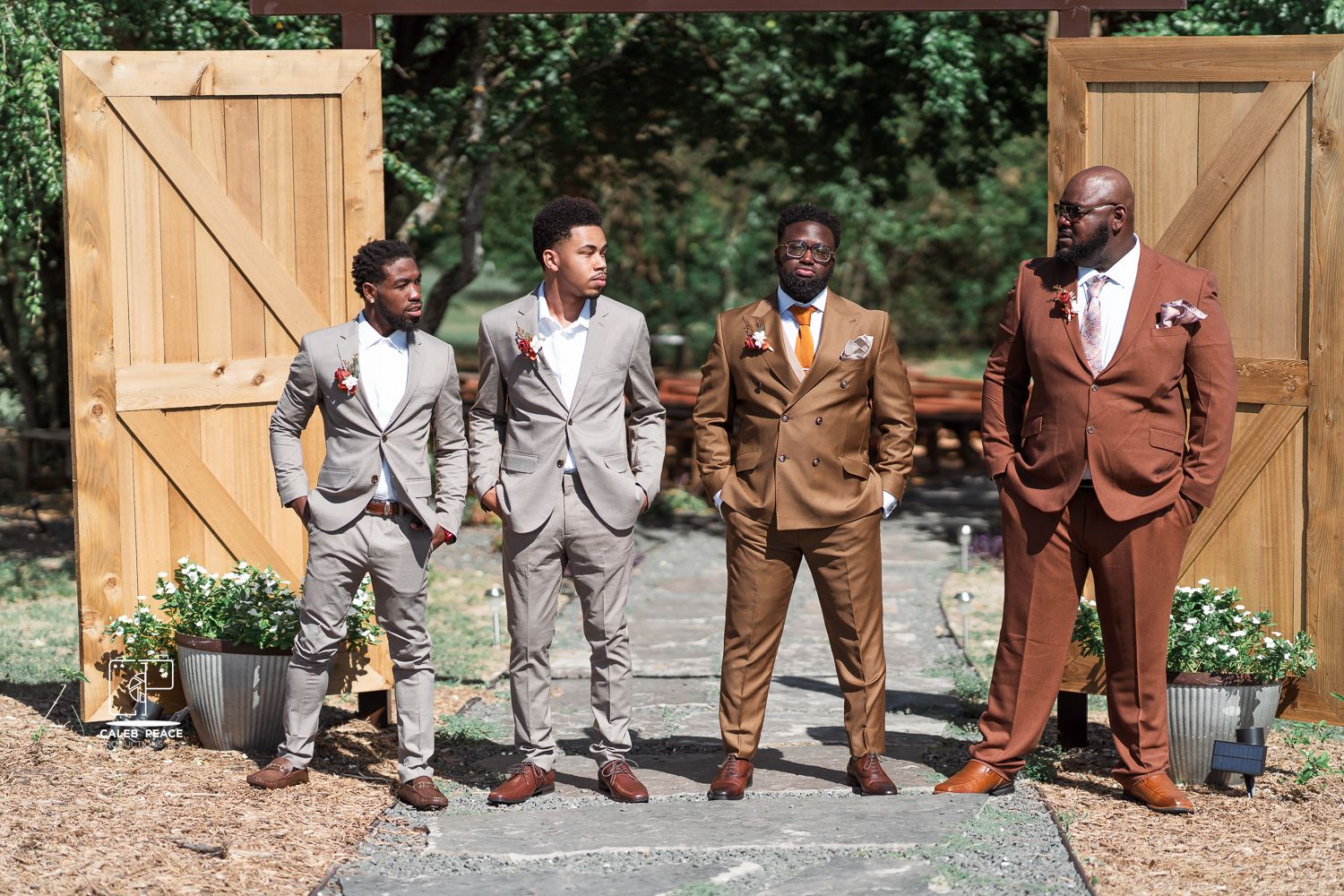 Four men in stylish suits pose together outdoors in front of wooden barn doors with potted plants.