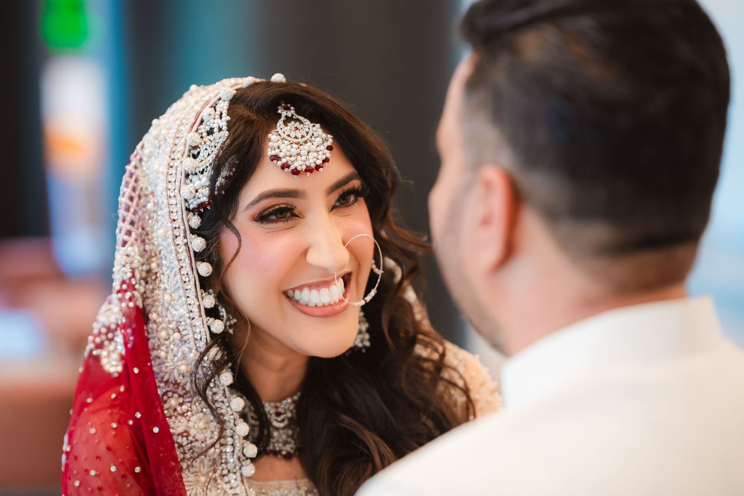 A traditional South Asian bride in an ornate red and white wedding dress smiles during her wedding ceremony.
