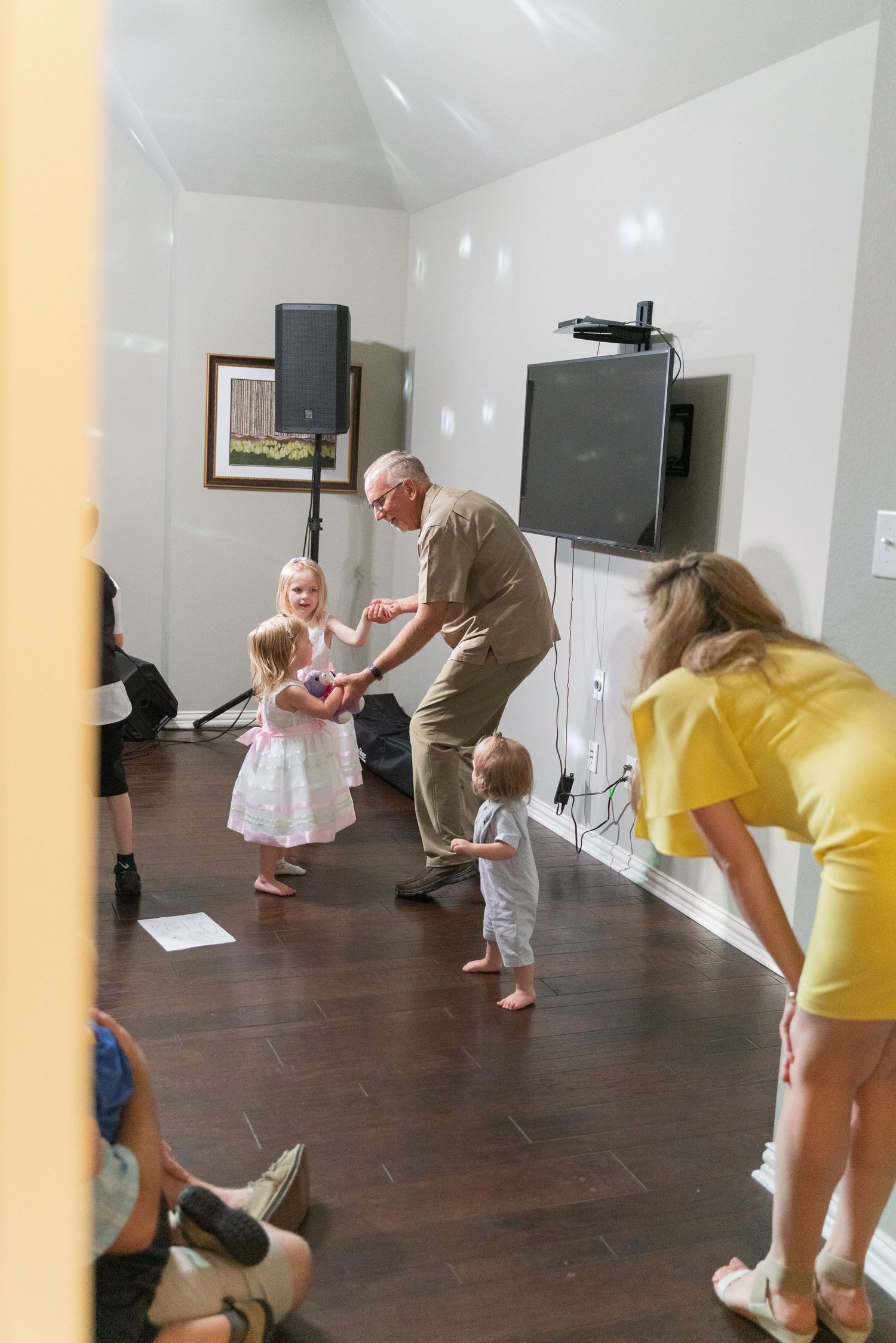 Children play and dance on a dark hardwood floor while adults in casual clothing watch.