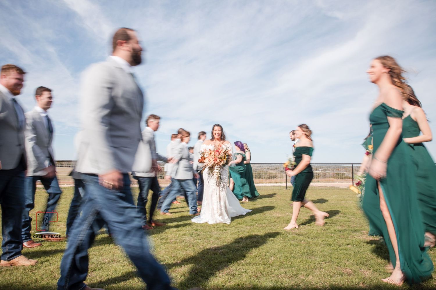Wedding party members run and dance joyfully across a grassy field on a sunny day.