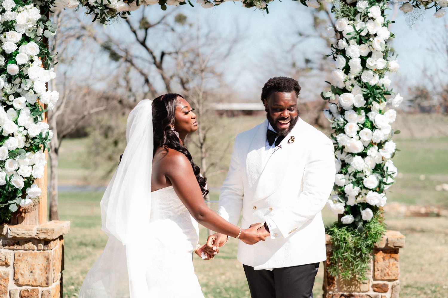A couple shares joyful moments under a floral arch during an outdoor wedding photoshoot in a natural setting.