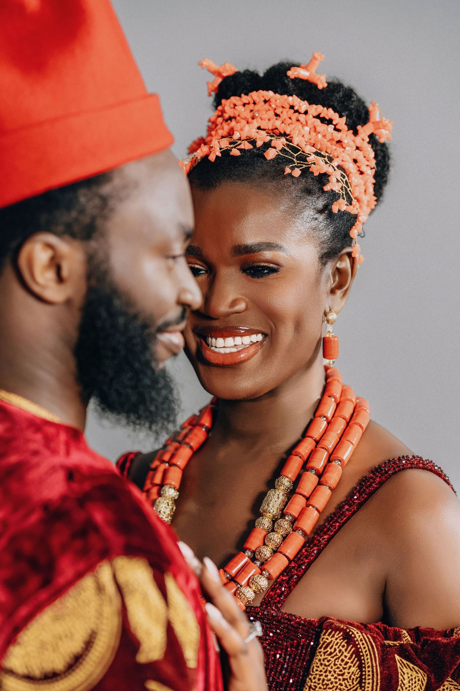 Traditional African wedding couple wearing red and gold attire with coral beaded jewelry in intimate portrait series.