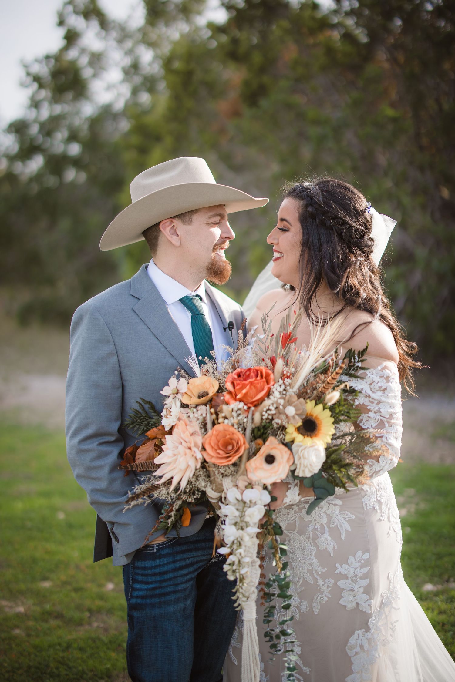 A romantic wedding couple embraces outdoors while holding a beautiful bouquet with orange roses and dried flowers.