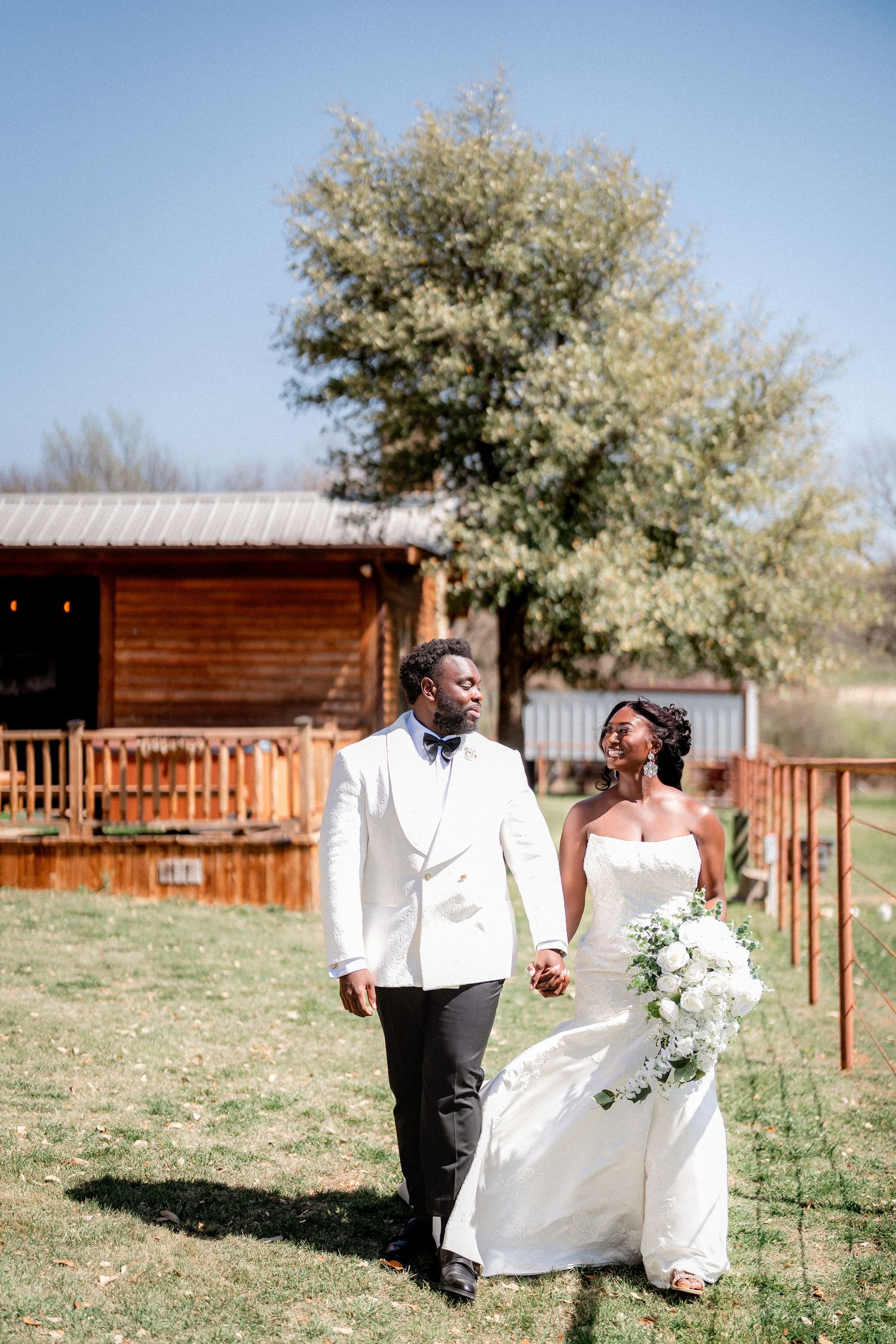 A wedding couple walks hand in hand on grass with a rustic cabin in the background.