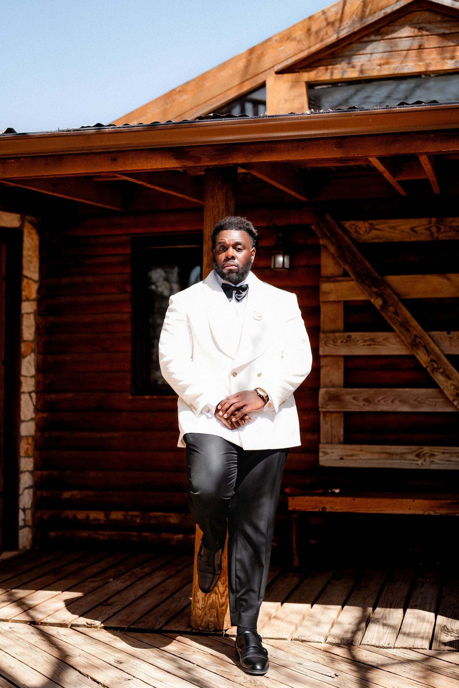 A figure in white shirt and black pants sits thoughtfully on wooden steps under a rustic overhang.