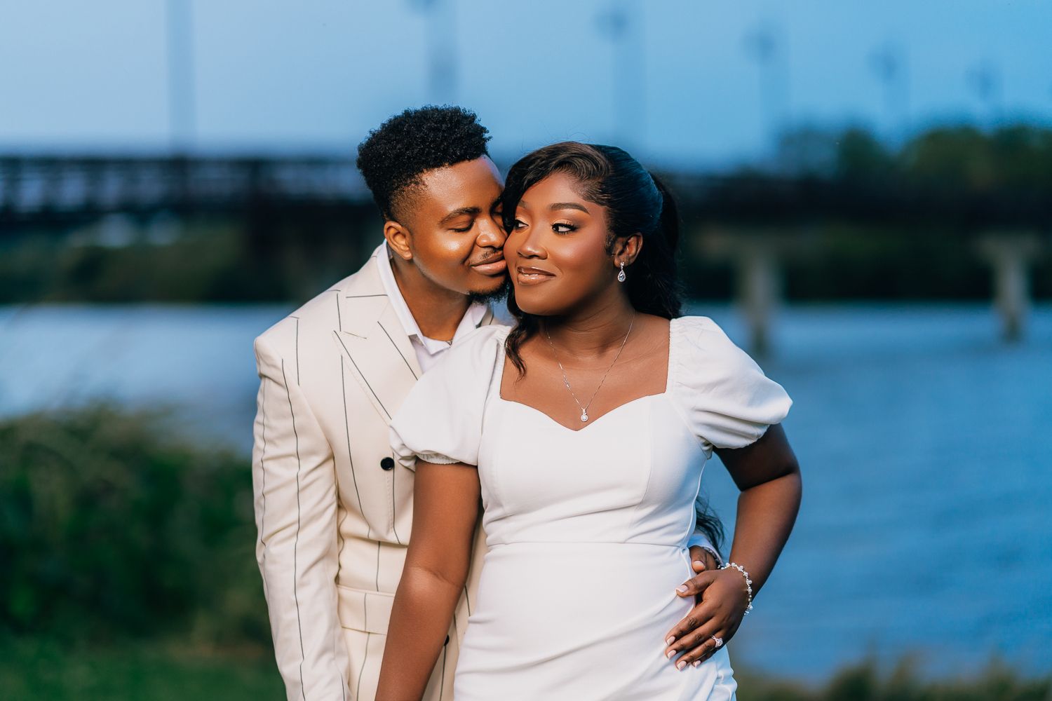 A couple dressed in white embraces by a waterfront setting during an engagement or wedding photoshoot.