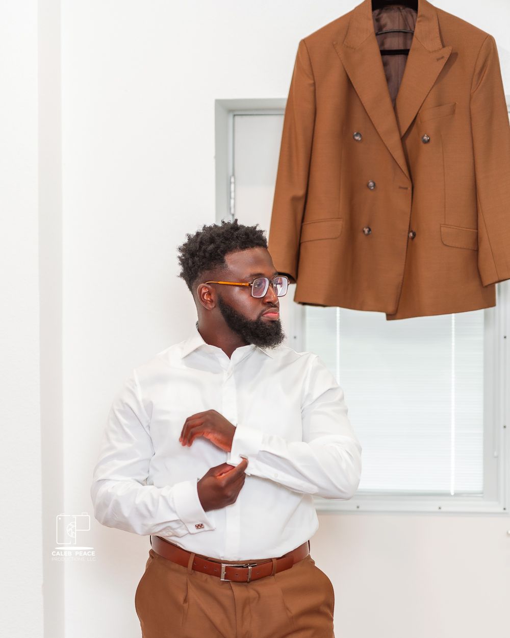 A well-dressed person in a white dress shirt and brown pants stands next to a hanging brown blazer jacket.