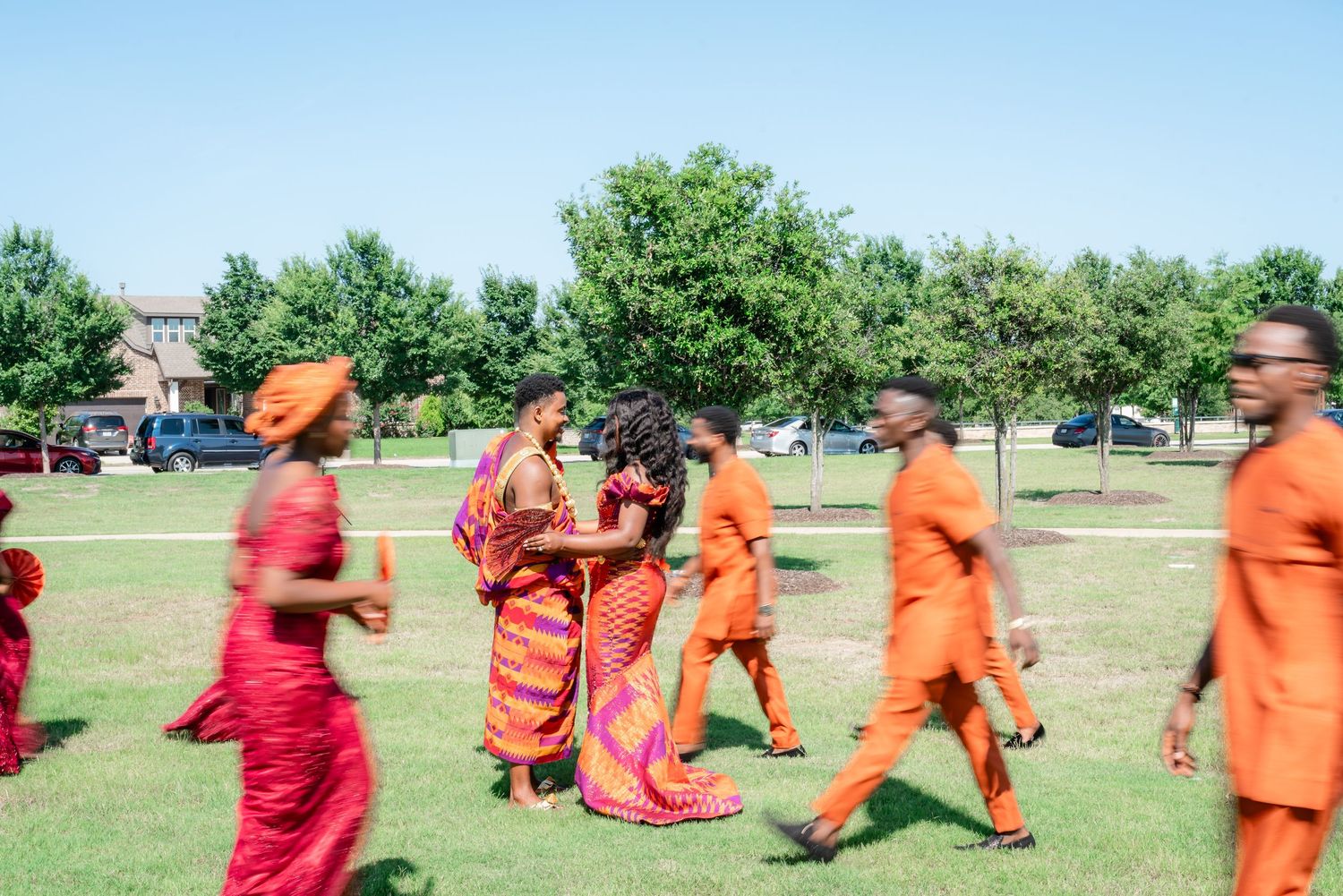 A group in vibrant orange and red traditional attire dancing together on a grassy field during a celebration.
