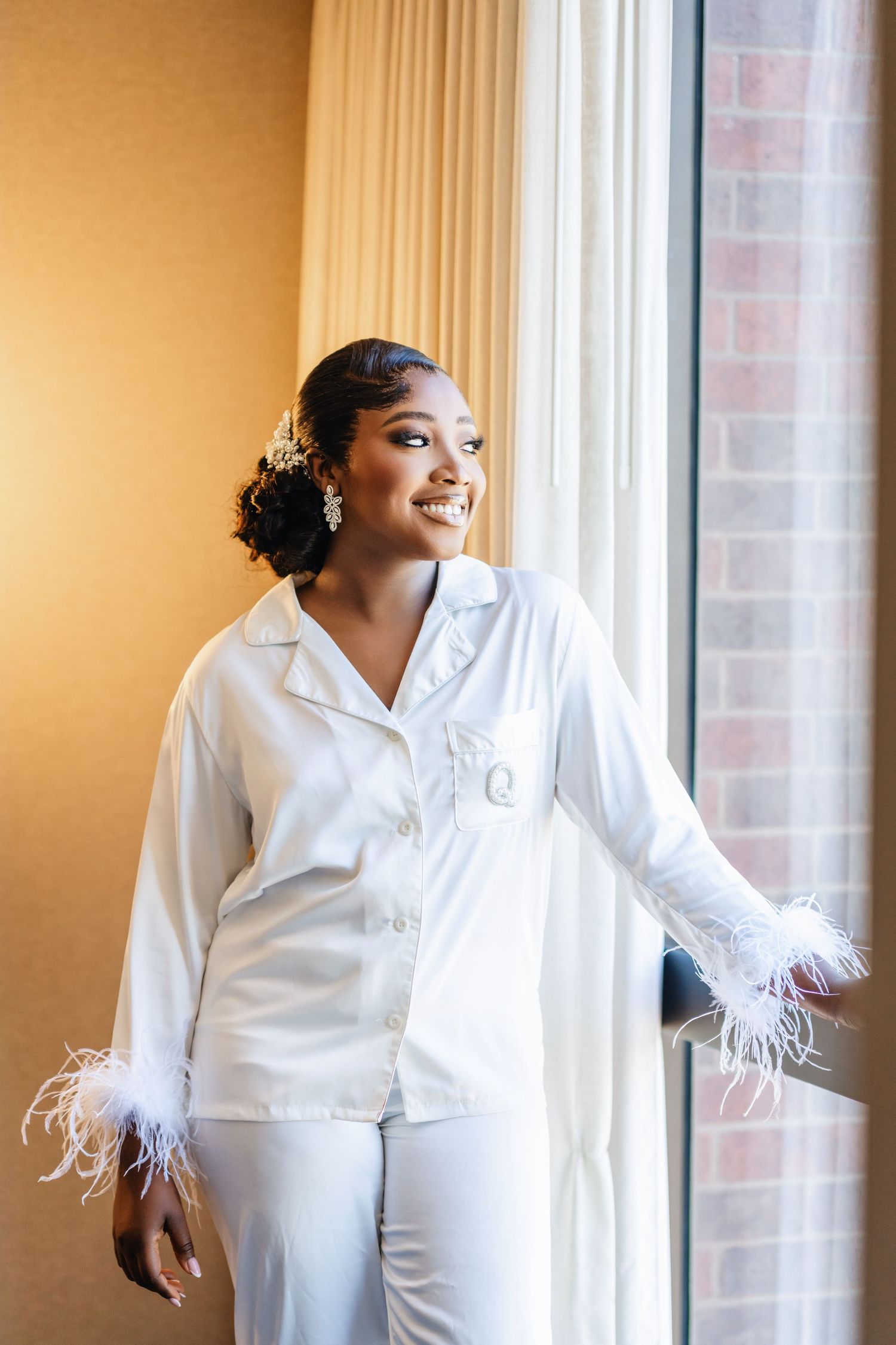A woman in a white satin robe with feather trim poses elegantly by a window.