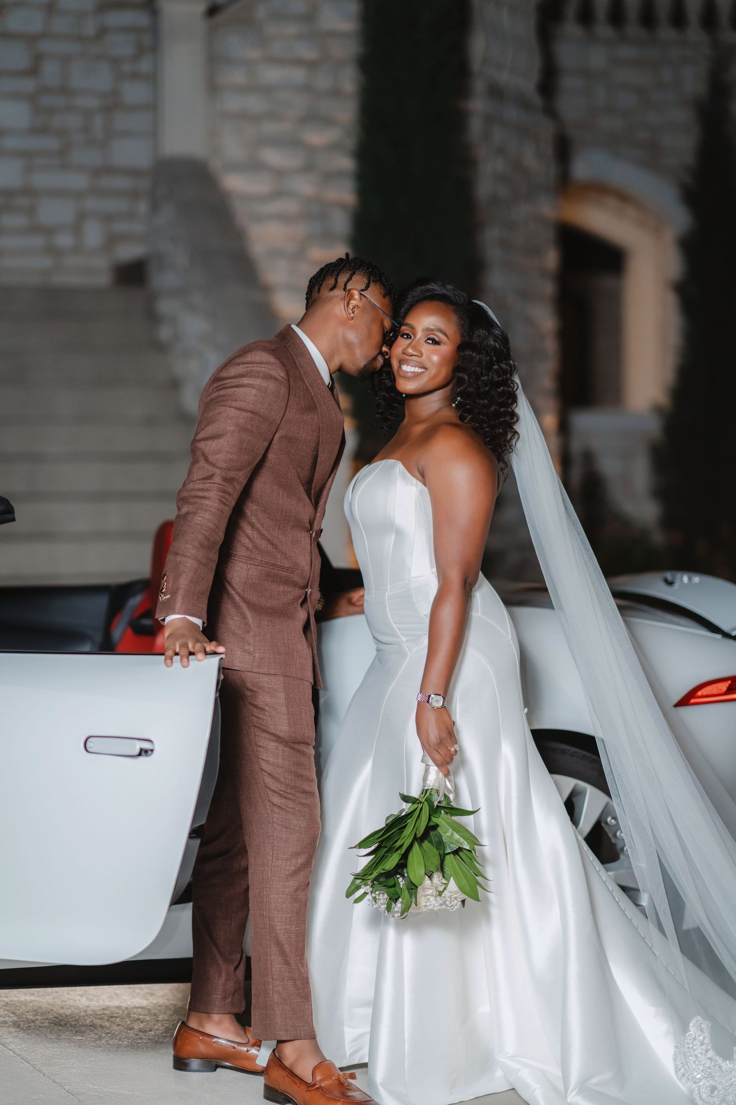 Newlyweds pose together near a white luxury car, with bride in flowing gown and groom in brown suit.
