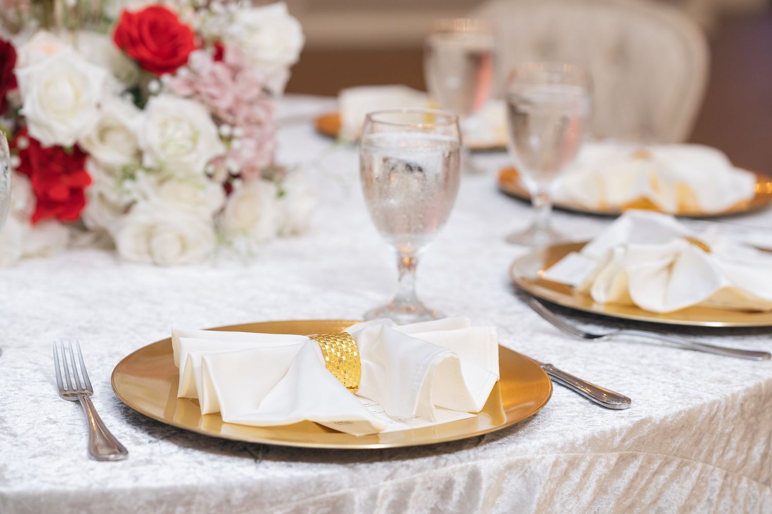 Elegant table setting with gold charger plates, white linens, and red and white floral centerpieces at a formal event.