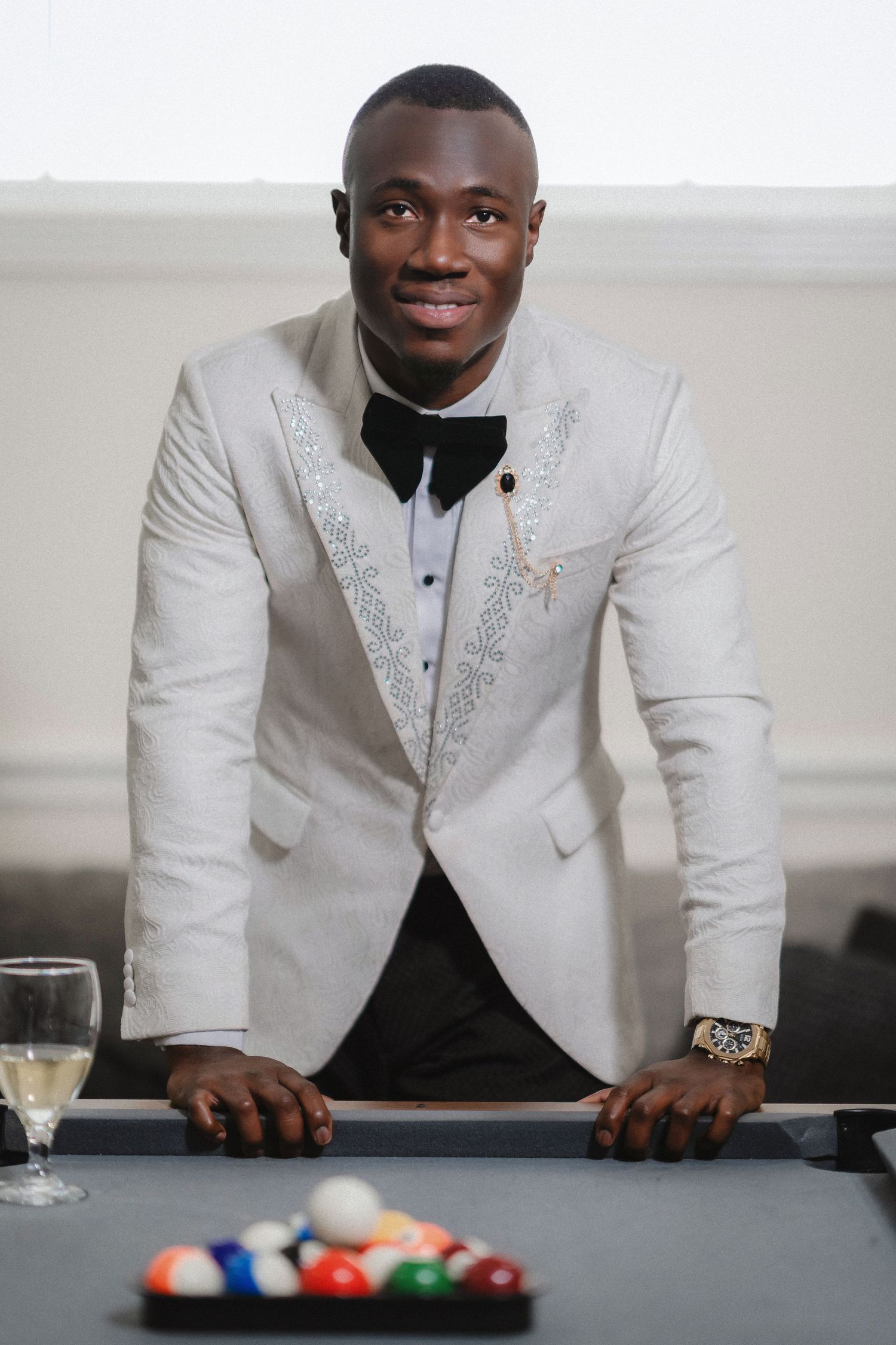 Dapper groom in embellished white dinner jacket stands at pool table with champagne and billiard balls.