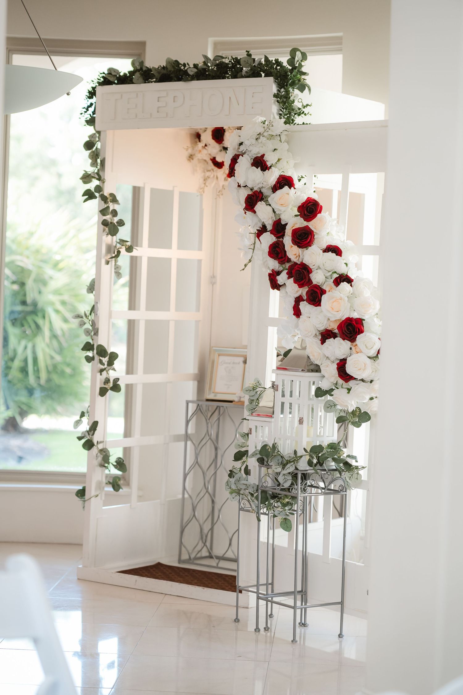 Elegant white wedding reception area decorated with red roses and greenery garlands.